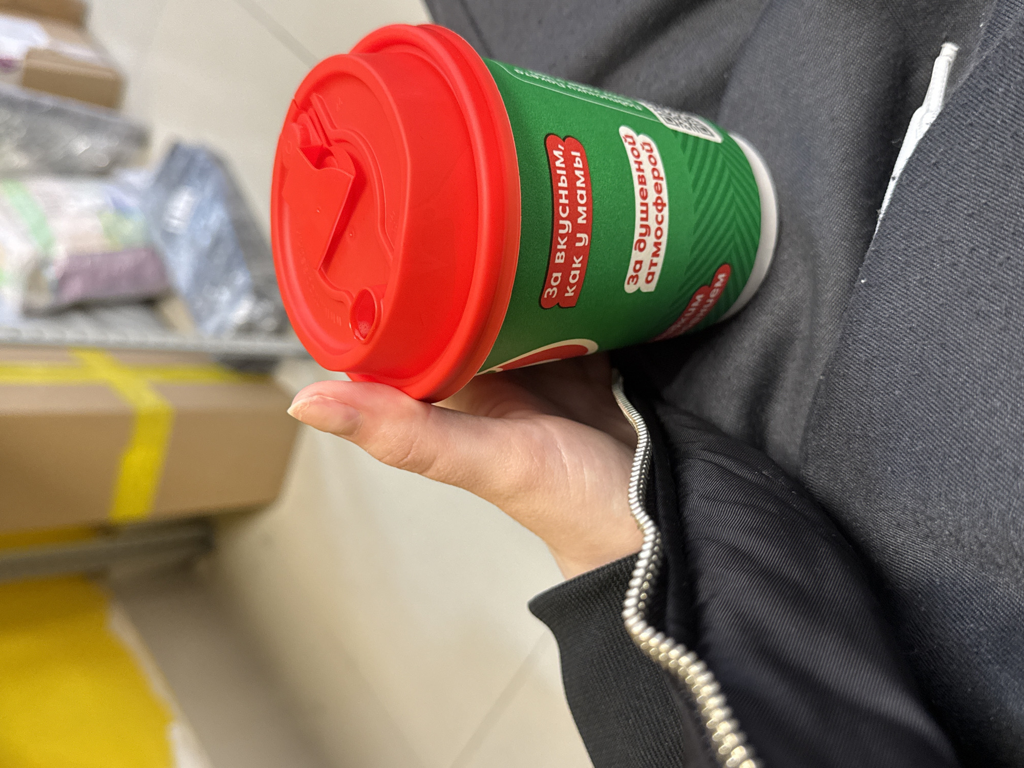 A person is holding a green and red coffee cup with Russian text on it in an indoor location. The person is wearing a dark jacket with a silver zipper. Boxes and other items are visible in the background. The location appears to be a shop in the city of Unknown, Unknown.