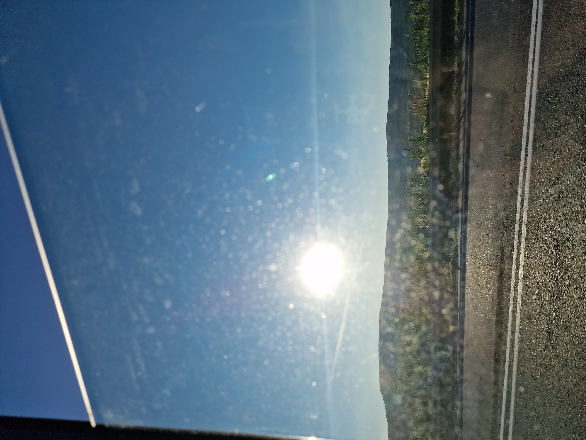 The image captures an outdoor scene from the perspective of looking out a vehicle's side window, rotated 90 degrees clockwise from its likely original landscape orientation. When viewed correctly, a bright blue sky dominates the upper portion, featuring a strong, brilliant sun causing significant lens flare and numerous bokeh effects, which appear to be dust particles or water droplets on the windowpane.
Below the sky, a rural landscape unfolds with a gentle horizon line revealing rolling hills or elevated terrain covered in green vegetation, indicative of fields or natural growth. On the far right, a dark asphalt road with two distinct parallel white lines is visible in the foreground, suggesting a highway or main road. The imperfections of the glass surface, including reflections and specks, are noticeable throughout the image. The scene depicts a clear, sunny day, likely during the mid-morning or afternoon, indicating travel through a rural area. No visible text is present. The setting is Tatarka, Russia.