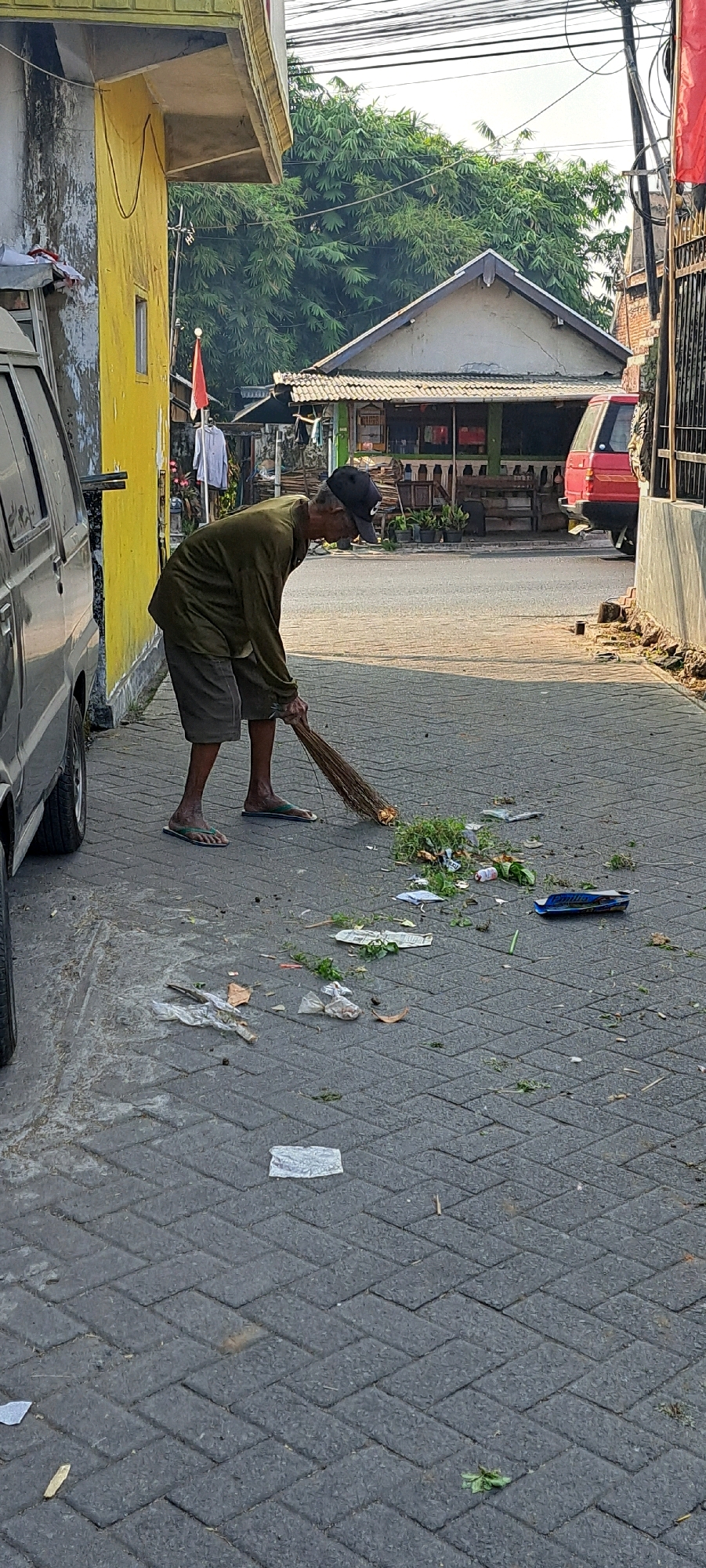 An elderly man wearing a dark cap, long-sleeved olive shirt, and shorts is sweeping debris on a paved walkway. He is using a broom made of natural fibers and is moving a pile of leaves and plastic waste to his right. The scene is set on a street with a cobblestone-like pattern, bordered by buildings. To the left, a portion of a dark-colored van is visible. In the background, there are structures with awnings, trees, and power lines, suggesting an urban environment. The presence of a small Indonesian flag on a pole and the architectural style of the buildings point to a location in Indonesia, possibly Candi Wates, Indonesia. The lighting suggests it is daytime, with shadows cast on the ground.