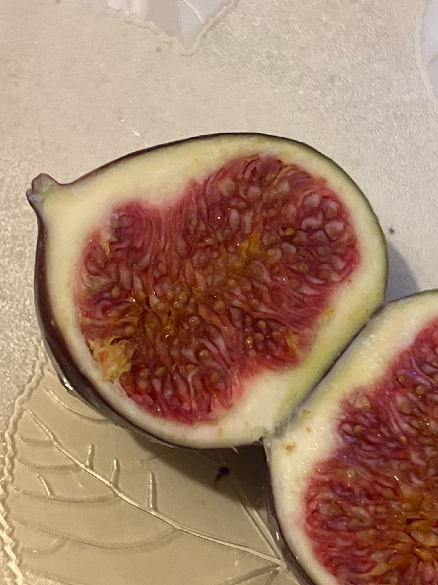 The image shows a close-up of two halves of a fig cut in half, displayed on a decorative glass plate with a leaf pattern. The figs have dark purple skin and vibrant pink flesh with visible seeds. The background is a textured, light-colored surface, possibly a tablecloth or countertop. There are no people or any other objects visible in the image. The lighting is soft and highlights the glossy texture of the figs. The scene is static and depicts a still life arrangement of fruit.