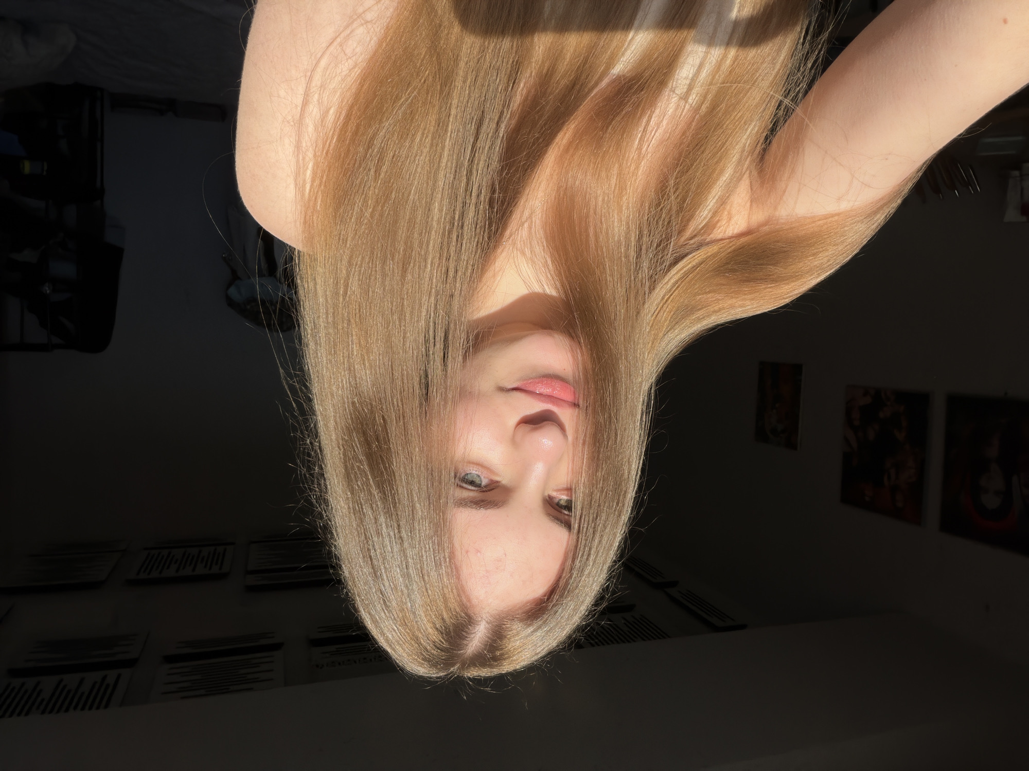 The image shows a young person with long, blonde hair, illuminated by natural light. Their face is angled upwards, with their eyes looking directly at the camera. The person has light-colored eyes, rosy cheeks, and pink lips. Their hair is parted in the middle and falls over their shoulders and down their back. Sunlight casts shadows across their face and hair, creating a dramatic effect.
The background is dark and out of focus, with some framed pictures visible on the wall. The overall impression is that of a casual selfie taken indoors, likely during the day given the strong sunlight. The location context of Dnipro, Ukraine, is noted but not visually evident in the image itself. There is no visible text in the image.