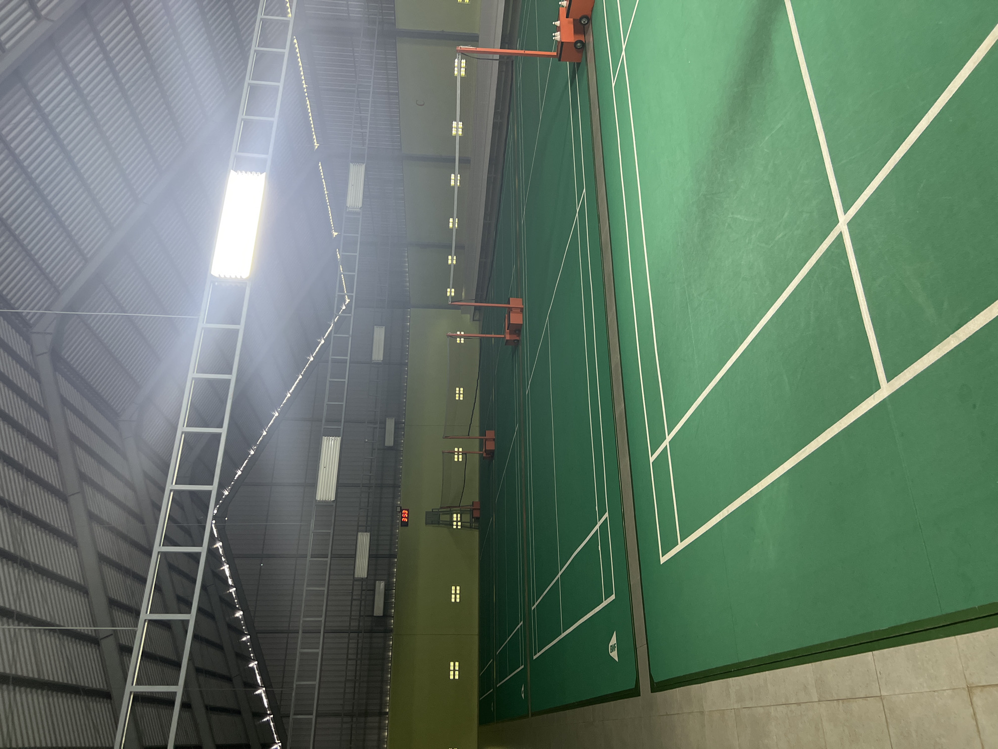 The image shows an indoor badminton court located in Kuta Selatan, Indonesia. The court is spacious with a green synthetic surface marked with white lines to delineate the playing areas. Two badminton nets are set up across the court. The walls are painted a light green, and there are rectangular light fixtures on the walls and ceiling, with one large light panel illuminating the center of the court from the ceiling. A digital scoreboard displaying "3:53" is visible in the background. The ceiling is constructed with corrugated metal sheeting supported by metal beams, typical of indoor sports facilities. There are no people visible in the image. The environment is clean and well-lit, suggesting the facility is ready for play.