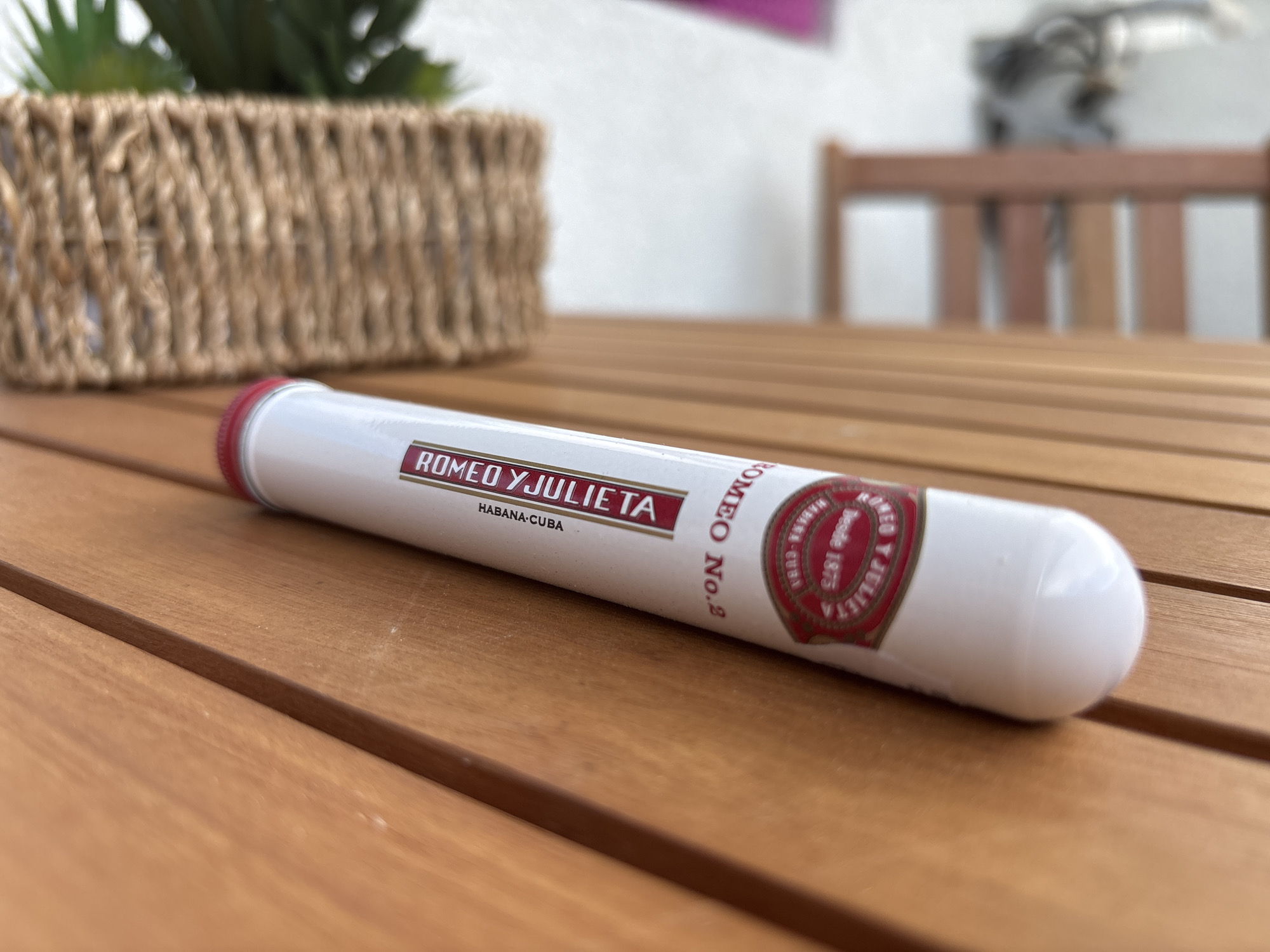 A close-up, low-angle shot shows a Romeo y Julieta No. 2 cigar in a white tube with a red cap resting horizontally on a wooden slatted table. The cigar is positioned diagonally from the bottom left to the upper right of the frame. The tube features red and gold branding that reads "ROMEO Y JULIETA" and "HABANA-CUBA". To the left of the cigar and out of focus, a portion of a woven basket with a plant is visible. In the background, also out of focus, is a portion of a wooden chair and a white wall. The lighting suggests an outdoor setting during the daytime.