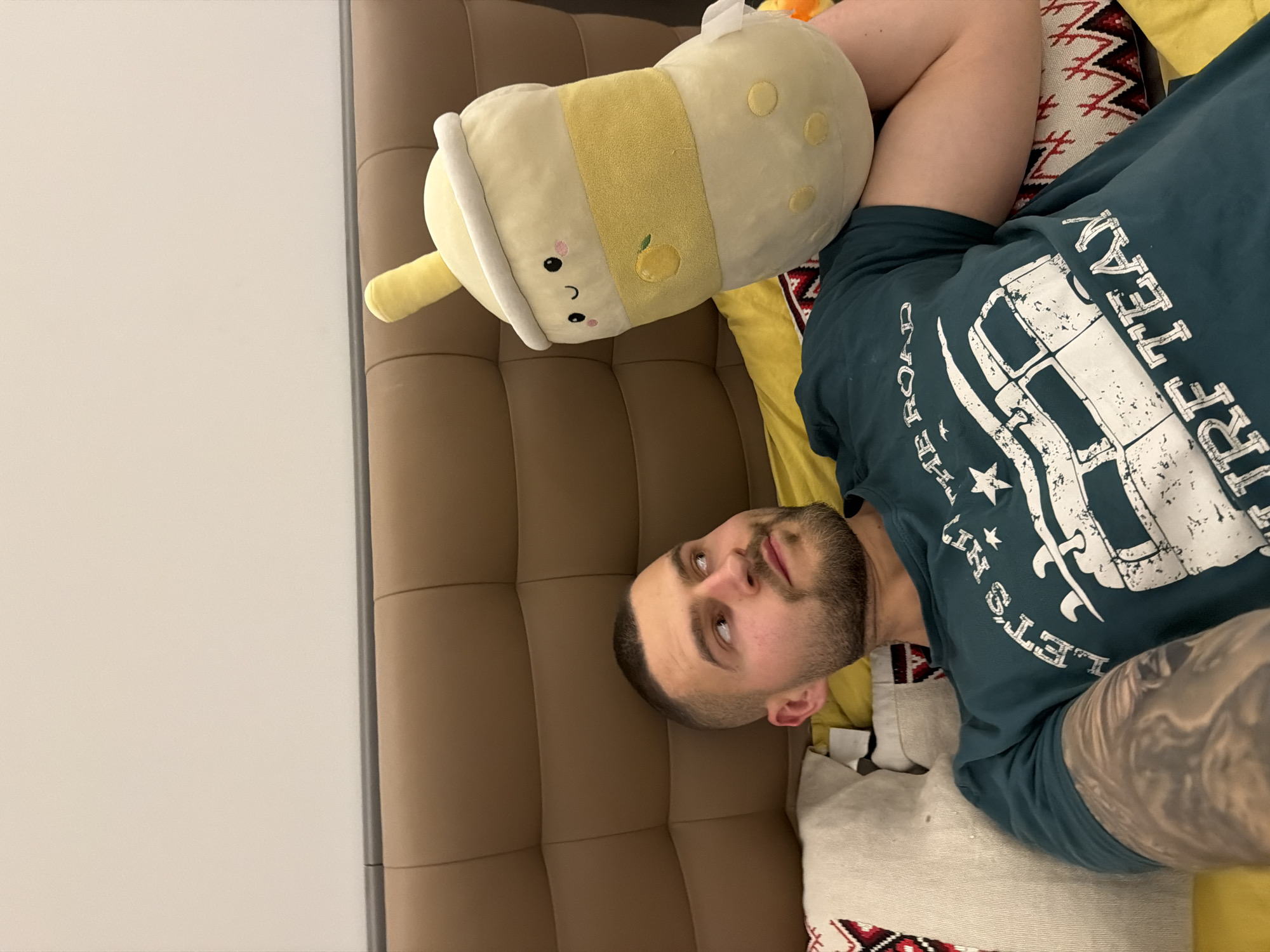 A man with short, dark hair and a beard is lying on his back on a brown, tufted surface, possibly a sofa or bed. He is wearing a dark green t-shirt with a white graphic design. His left arm is visible and shows a tattoo. He is looking at the camera. On his right side, he has a large, light yellow plush toy resembling a bubble tea drink. There is a yellow and red patterned cushion behind him. The location cannot be determined based on the image.