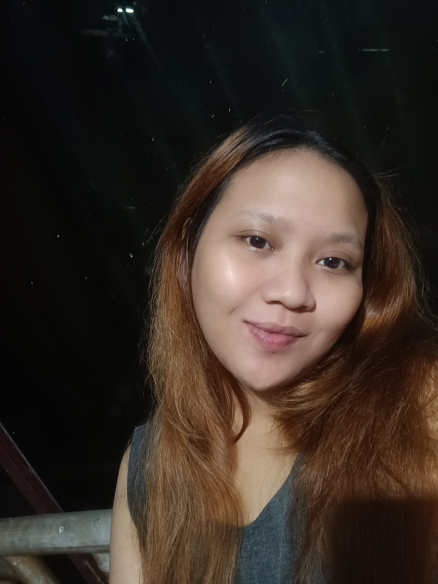 A close-up, selfie-style photograph shows a young woman with long, reddish-brown hair. She is looking directly at the camera with a slight smile. Her skin is fair, and she has dark eyes. She is wearing a gray sleeveless top. The background is dark, suggesting it is nighttime, with some faint lights visible, possibly from distant streetlights or stars. A portion of a metal railing and wooden structure is visible in the lower left corner, suggesting an outdoor setting, possibly a balcony or porch. The overall mood is casual and relaxed. The location is identified as Depok, Indonesia, and the time of day is likely evening or night. There is no visible text in the image.
