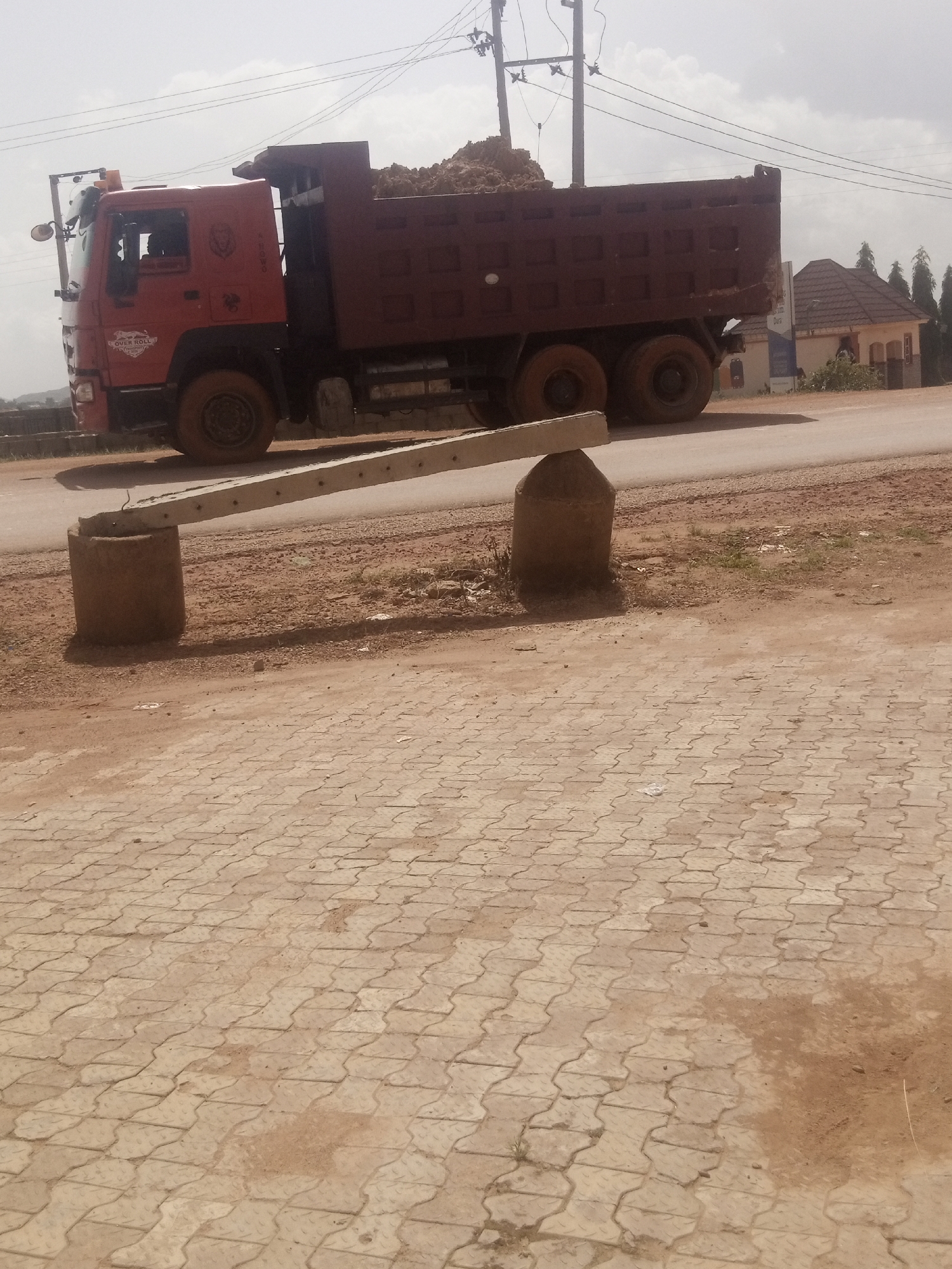 The image shows a dump truck filled with dirt, parked on a road. The truck is red and appears to be in motion or just stopped. It has multiple wheels and a large bed for carrying materials. In the foreground, there are two concrete posts supporting a tilted concrete beam, which could be part of a barrier or structure. The ground is a mix of dirt and paved interlocking bricks. In the background, there are some buildings with tiled roofs and trees, suggesting an urban or semi-urban environment. Overhead, power lines are visible against a cloudy sky. The overall scene appears to be during the day, with the lighting suggesting it could be late morning or early afternoon. The weather seems overcast or hazy. There is some text on the truck, including "OVER ROLL" and a logo with a lion. The scene suggests a construction or transportation activity, possibly related to road work or earthmoving.