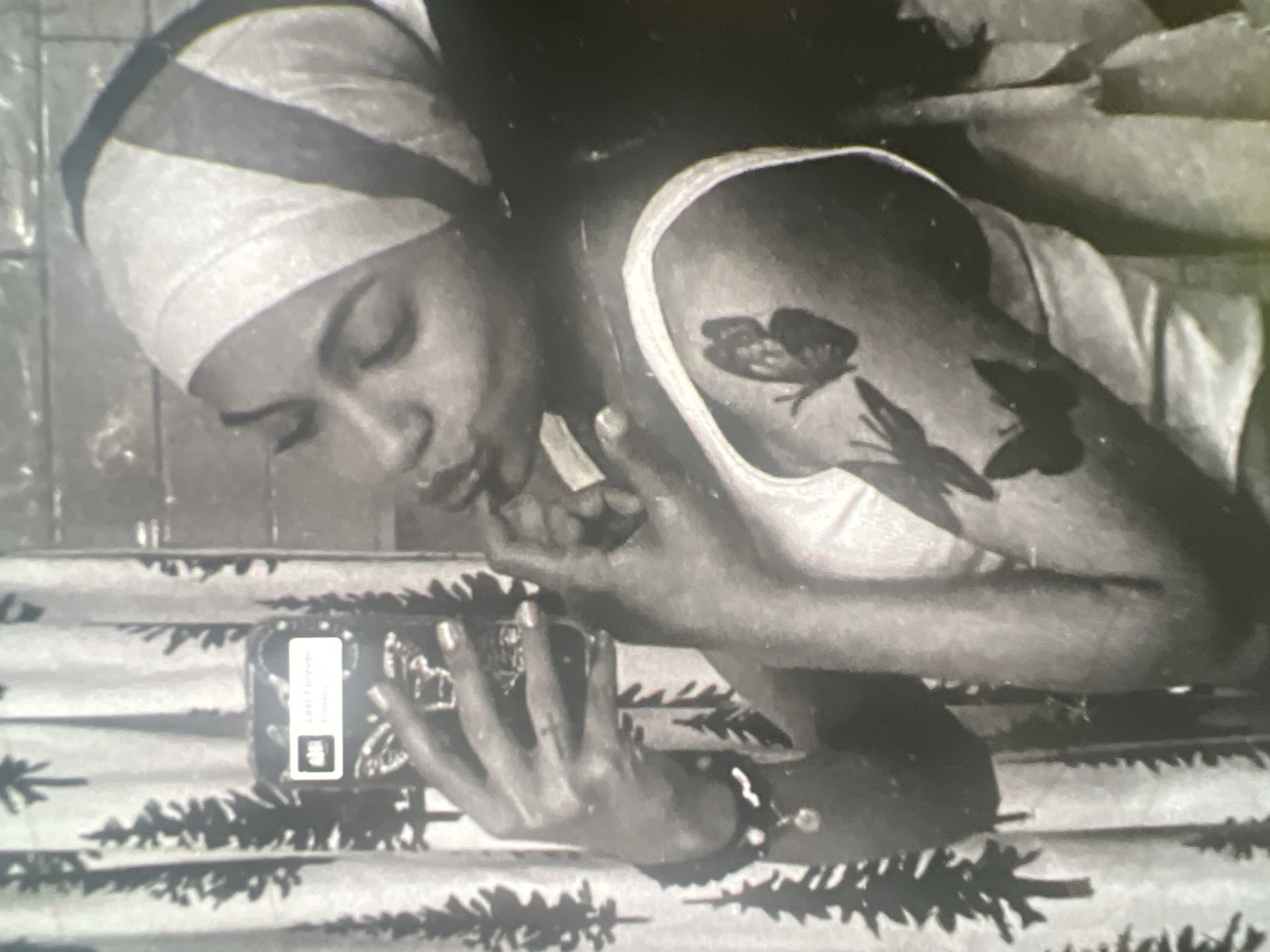 A black and white, medium-shot, selfie-style photograph shows a young Black woman in profile, looking into her phone as she takes a picture. She is wearing a light-colored tank top and a headscarf with dark stripes. She has a tattoo of three butterflies on her left arm. Her right hand is raised to her face, with her fingers touching her lips. In her left hand, she holds a phone with a butterfly design on the case. The background is a shower curtain with a pattern of trees. The image appears to have been taken in a bathroom. The exact location in the city of Boone, United States of America, is not discernible from the image.