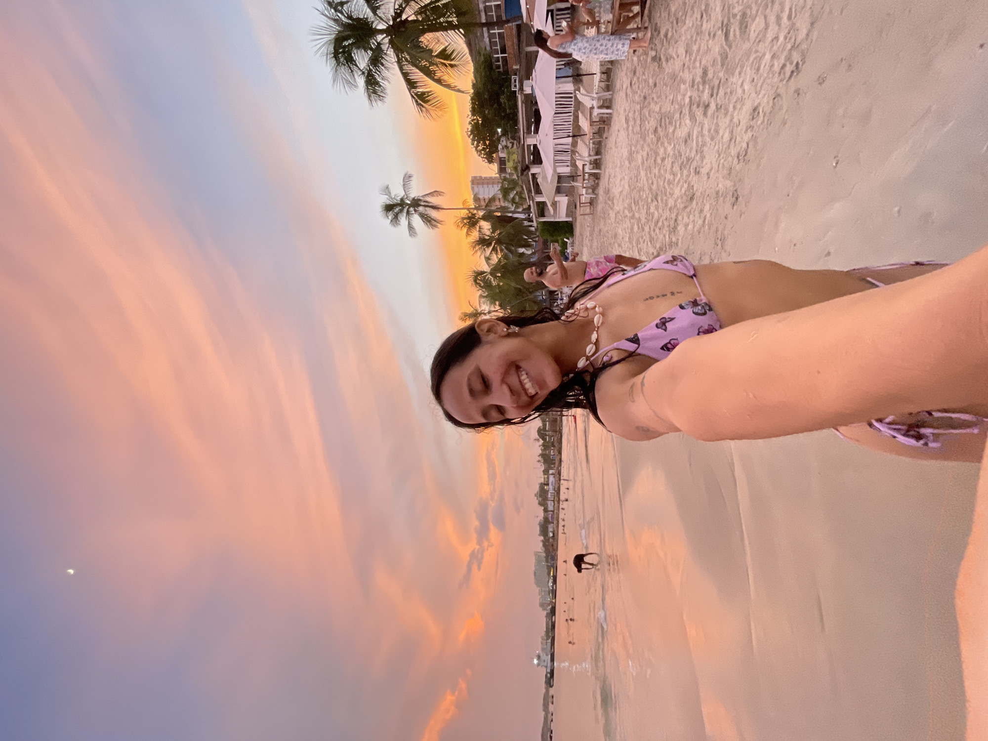 A woman wearing a purple butterfly-patterned bikini smiles at the camera while taking a selfie on a beach at sunset in Pampatar, Venezuela. The sky is a gradient of pink and orange, with a sliver of the moon visible. In the background, palm trees line the sandy shore, and several people are scattered along the beach and in the shallow water. Buildings are visible in the distance, suggesting the beach is near a town. A dog is in the water near the woman. The scene depicts a peaceful evening at the beach.