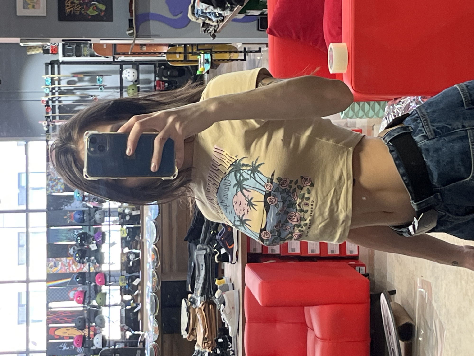 A person is taking a selfie in a store. The person is wearing a crop top, jeans, and a black belt. The store appears to be selling skateboards, helmets, and shoes. The interior includes red seating and visible window views of a cityscape. The location is possibly in Krasnoyarsk, Russia.