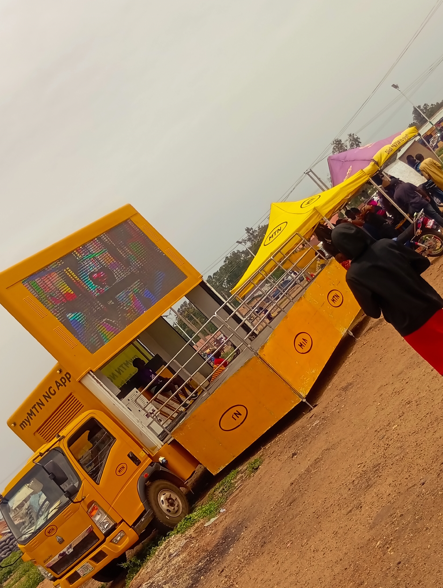 The image shows an MTN (Mobile Telephone Network) truck with "myMTN NG App" written on it, parked on a dirt ground in an outdoor area. The truck is yellow, with MTN logos and lettering on the side. There are also tents with the MTN logo in the background and some people standing around. The setting appears to be in Bukuru, Nigeria, as evidenced by the signage and the overall environment.