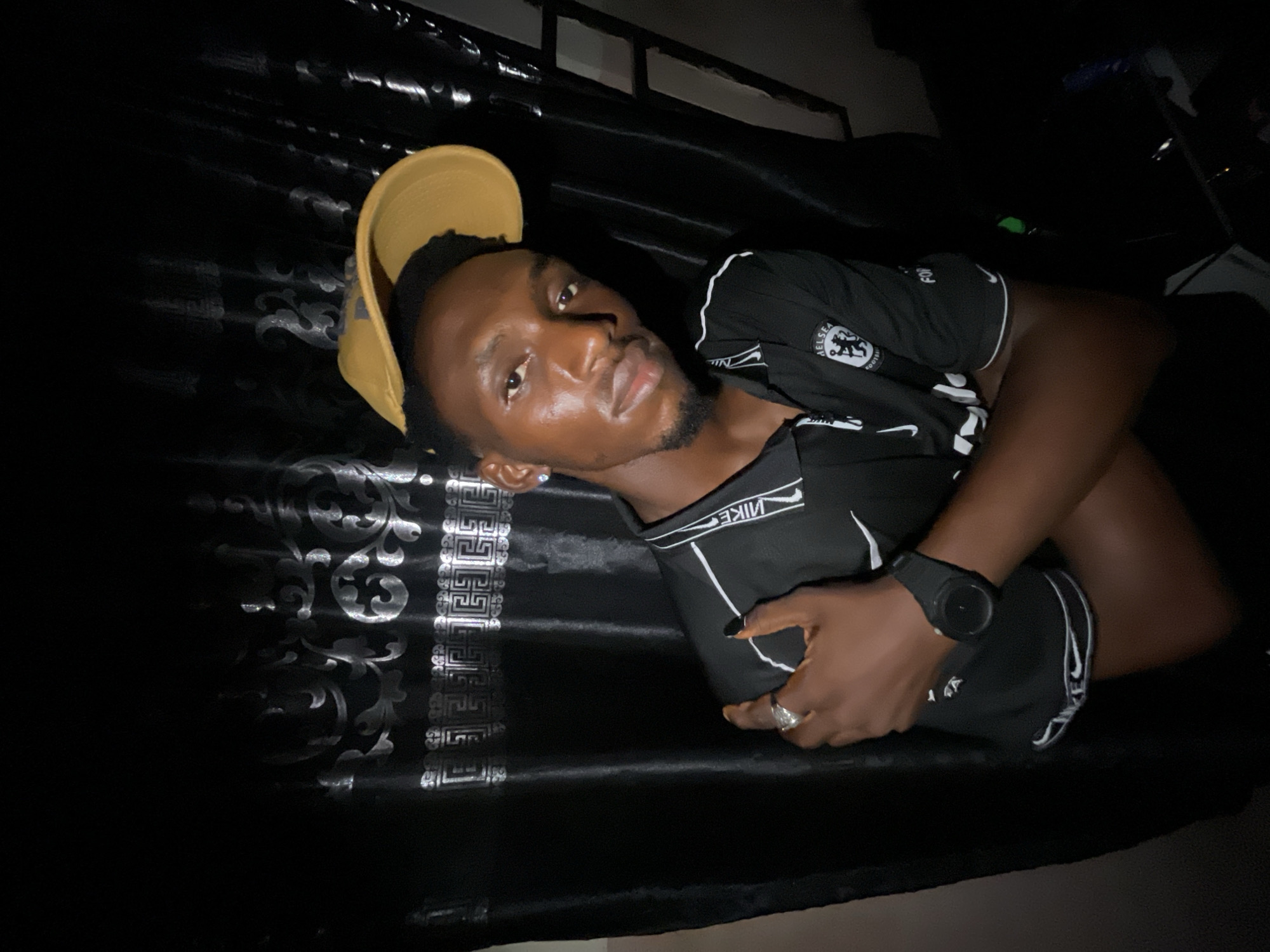 A young Black man is reclining on a dark, patterned surface. He is wearing a black Chelsea Football Club jersey and Nike shorts, also black. He is also wearing a gold-colored baseball cap and a black wristwatch on his left wrist. A silver ring is visible on his left hand. The photograph was taken indoors. It is too dark to pinpoint the exact location in Lokoja, Nigeria.