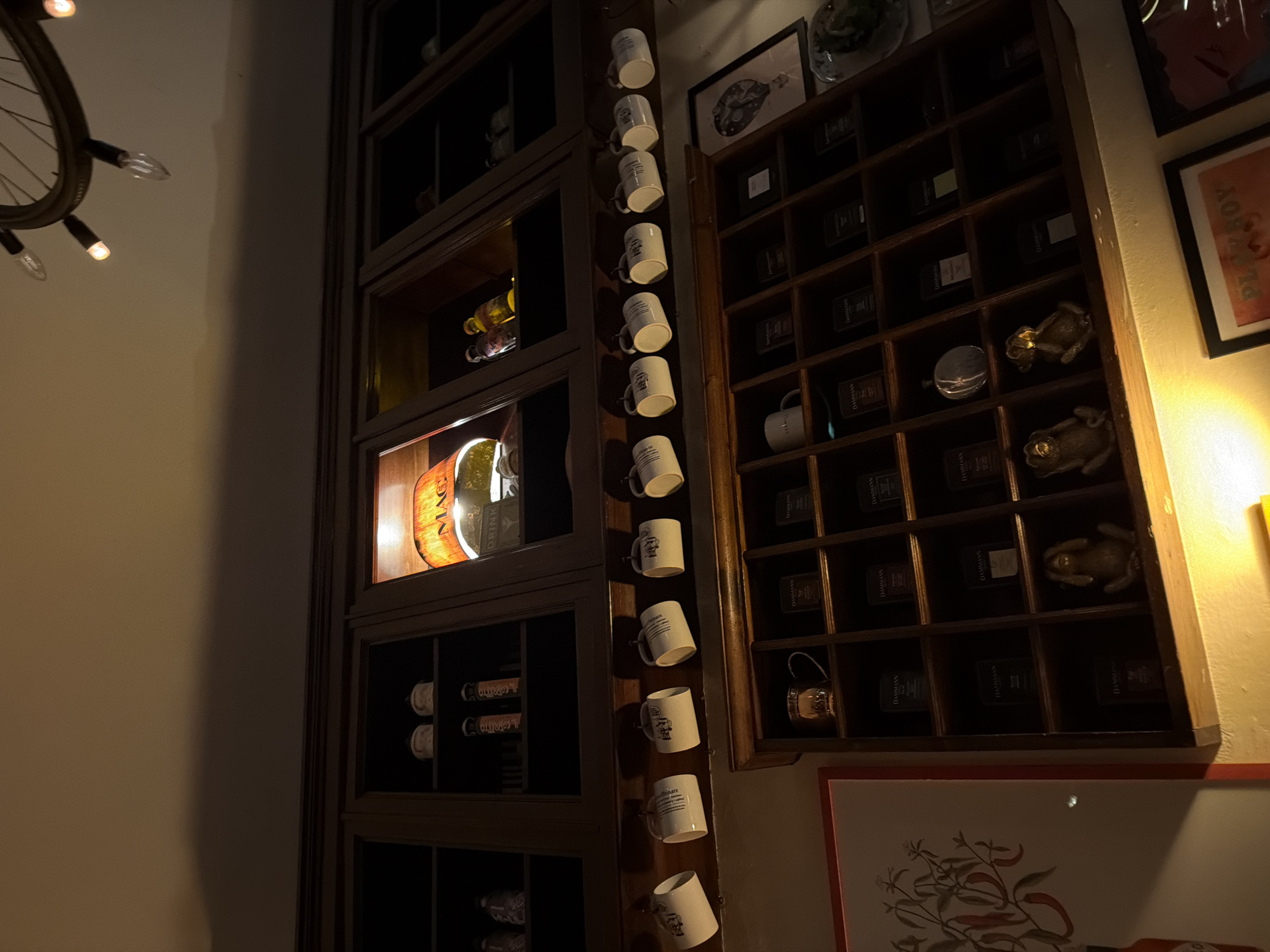 Here is a concise, forensic description of the image:
**Scene:** An indoor setting featuring two large, dark wood shelves.
**Objects:** The left shelf appears to be a wine rack displaying bottles of wine and an illuminated logo (possibly a brand or establishment name). Adjacent to it, hanging vertically, are a series of white ceramic mugs stacked atop one another, seemingly acting as decorative elements. The right shelf consists of a display cabinet with small compartments, each housing what appear to be tea boxes and decorative frog figurines.
**Environment:** The lighting is dim, creating a somewhat somber ambiance. The background is a cream-colored wall. A partially visible bicycle-wheel chandelier is hanging from the ceiling.
**Inferred Location:** The presence of tea boxes with the brand "DAMMANN" suggests a possible connection to the Dammann Frères tea company, which has stores in Milan. Given the combination of wine and tea, this could potentially be a high-end cafe, bistro, or restaurant in Milan.