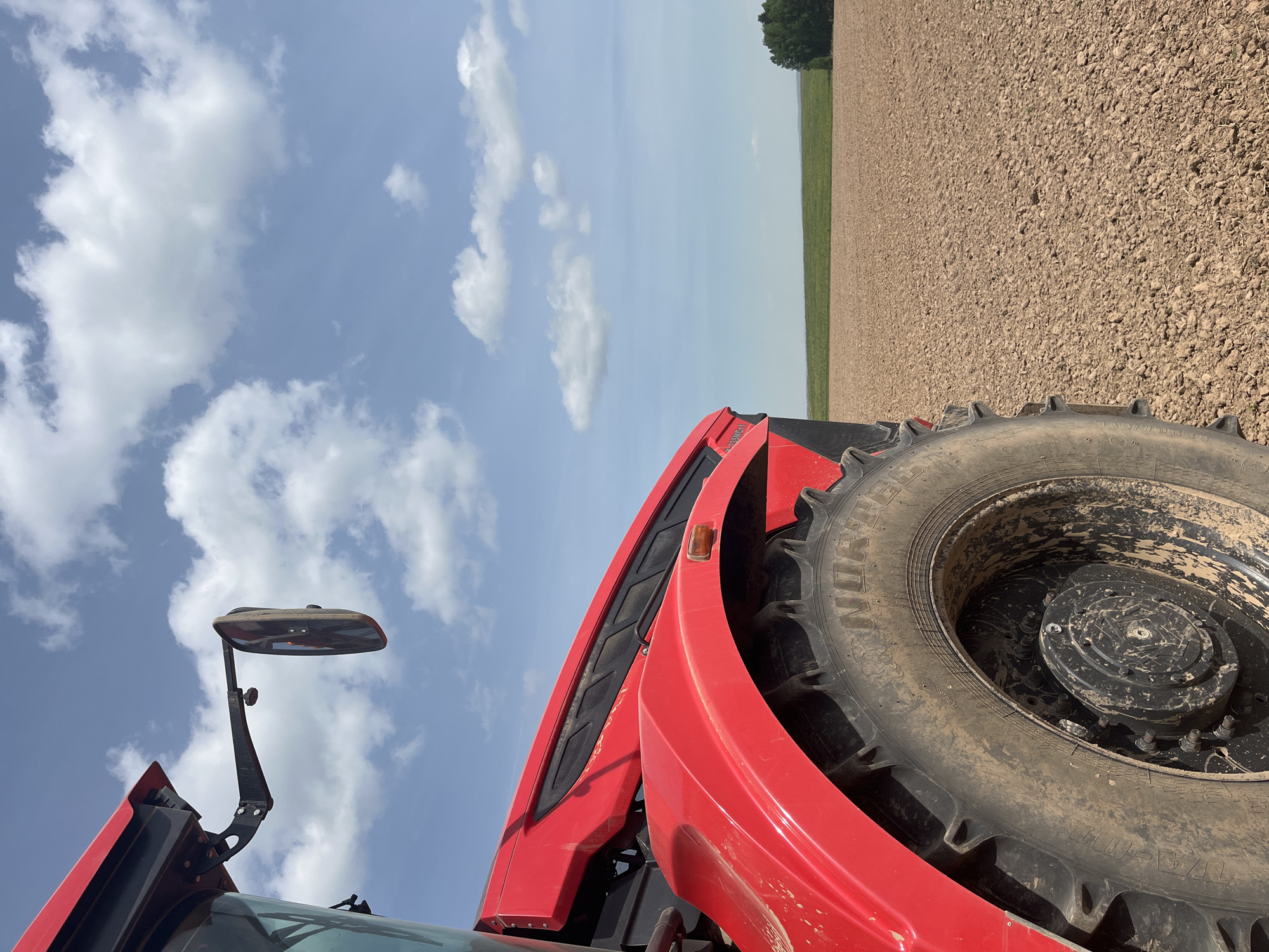 A low-angle shot depicts the front of a red tractor in a field. The tractor's large, muddy tire is prominently visible. The field appears plowed with exposed soil. A distant treeline marks the horizon. Above, a blue sky is dotted with scattered white clouds. The location is Unknown, Unknown.