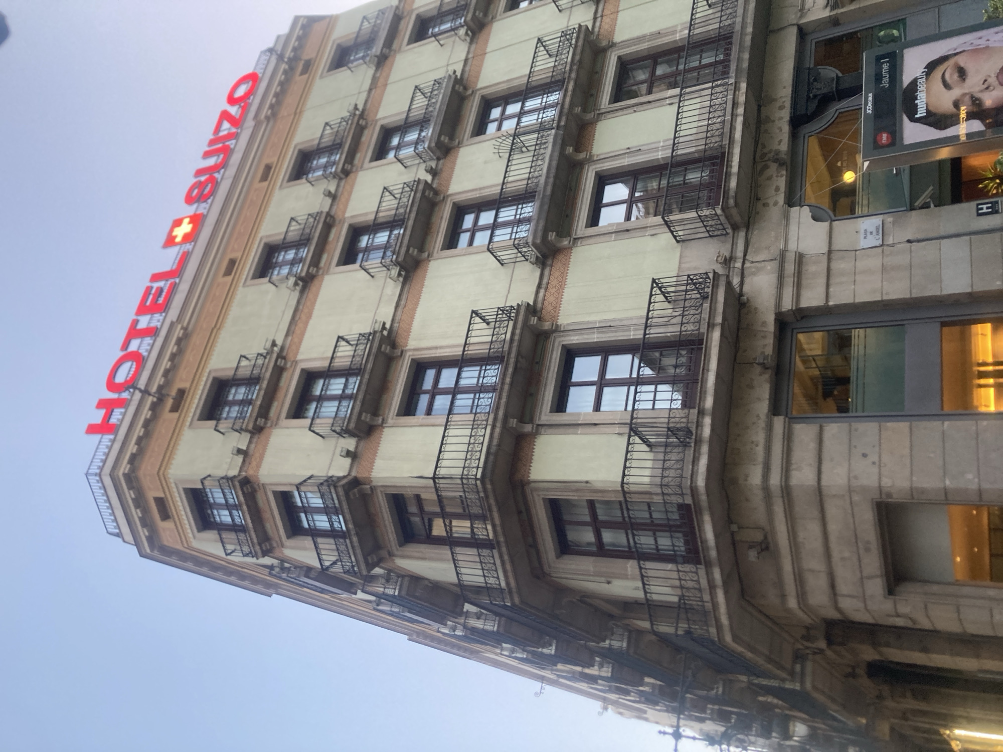 This is a low-angle, full shot of the corner of a building in Barcelona, Spain, under a clear sky. The building is tall with multiple stories featuring balconies with black wrought-iron railings. The facade is a pale yellow with decorative orange bands. Red neon lettering spelling "HOTEL SUIZO" with a red Swiss cross in the middle, is prominently displayed across the top of the building. On the ground level, large windows reveal an interior with warm lighting. A street sign reads "PLACA DE L'ANGEL" and an advertisement for "hudabeauty" is visible. There are no people visible in the image.