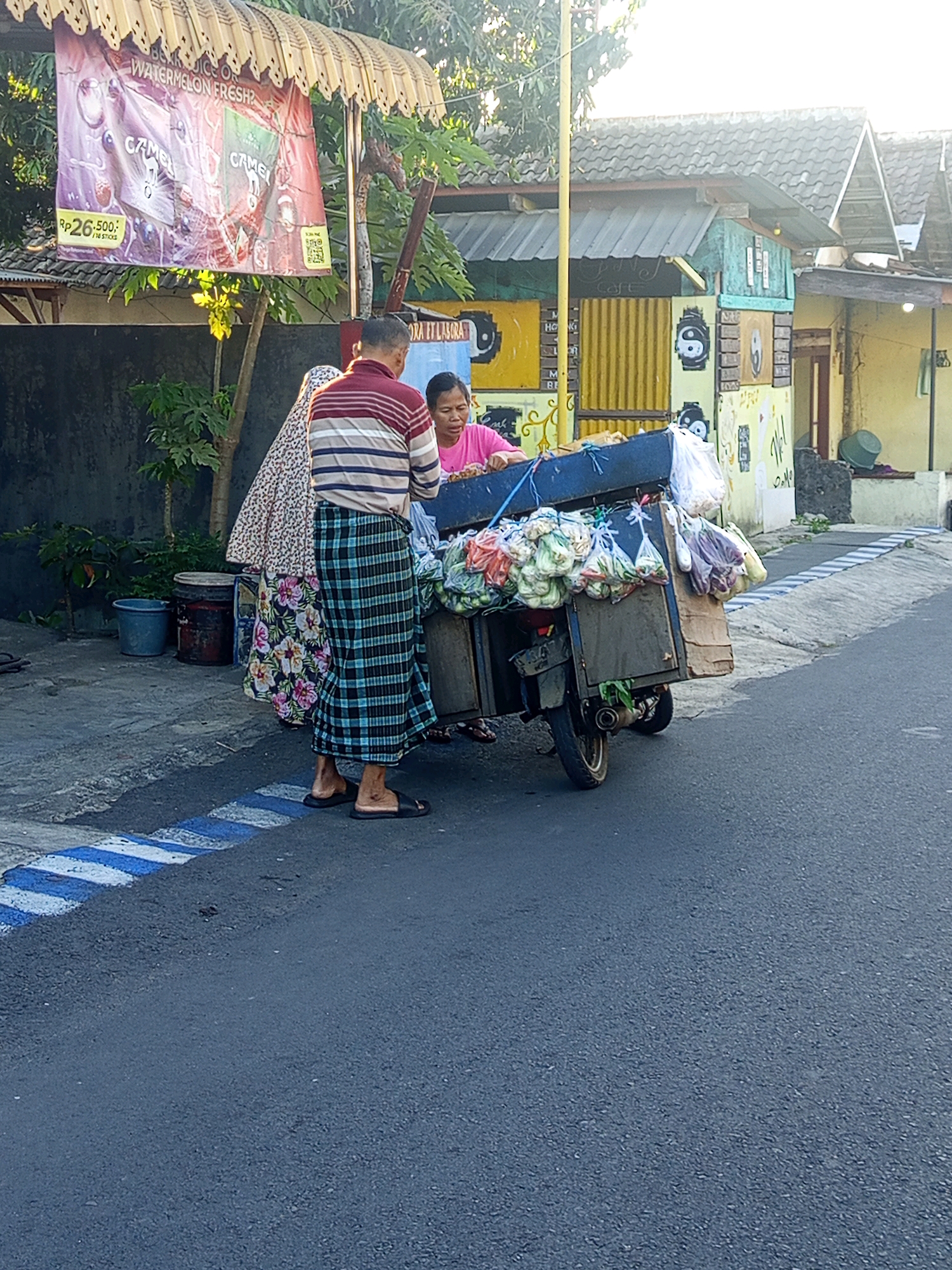 In Mulyorejo, Indonesia, a street vendor is engaged in commerce. A man, wearing a striped shirt and a patterned sarong, stands near a motorcycle with a sidecar loaded with produce. He is facing away from the camera and appears to be interacting with a woman in a pink shirt who is also near the motorcycle. A third person, partially obscured, stands behind the man, also wearing a patterned sarong. The motorcycle is parked on an asphalt road with blue and white painted markings on the side. In the background, a building with a corrugated metal roof and colorful decorations is visible, along with trees and a banner advertising "WATERMELON FRESH?" with "RP 26,500" displayed prominently. The scene appears to be in a residential or commercial area.
