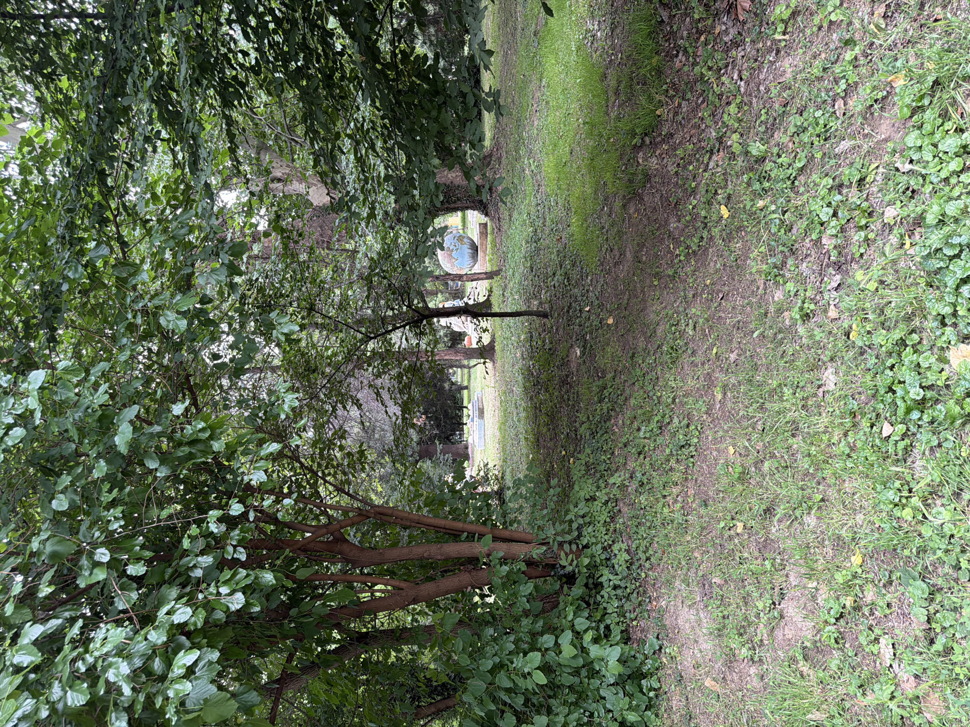 The image depicts an outdoor scene in a park-like setting. In the foreground, lush green foliage and grass dominate, with several trees and shrubs. The trunks of the trees in the foreground are a reddish-brown color. Further into the background, there is a cleared grassy area. In the middle ground, there is a large, spherical sculpture that resembles a globe, painted in blues and greens. Behind the globe sculpture, there appears to be a shallow, rectangular water feature or pool with some decorative elements, and a few indistinct figures or sculptures. The overall impression is a tranquil, natural environment with artistic installations. This scene is located in the City Park of Pazardzhik, Bulgaria.