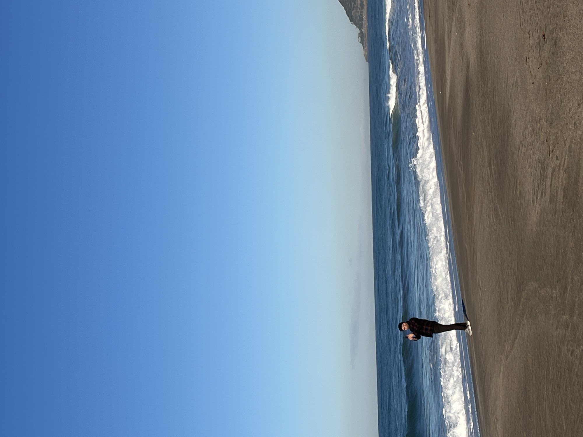 A wide-angle shot of a beach featuring a man in dark clothing and white shoes standing at the shoreline. Waves are breaking on the beach. The sky is clear blue. There is a headland or mountain visible in the upper-right corner of the frame. The location is Alanya, Turkey.