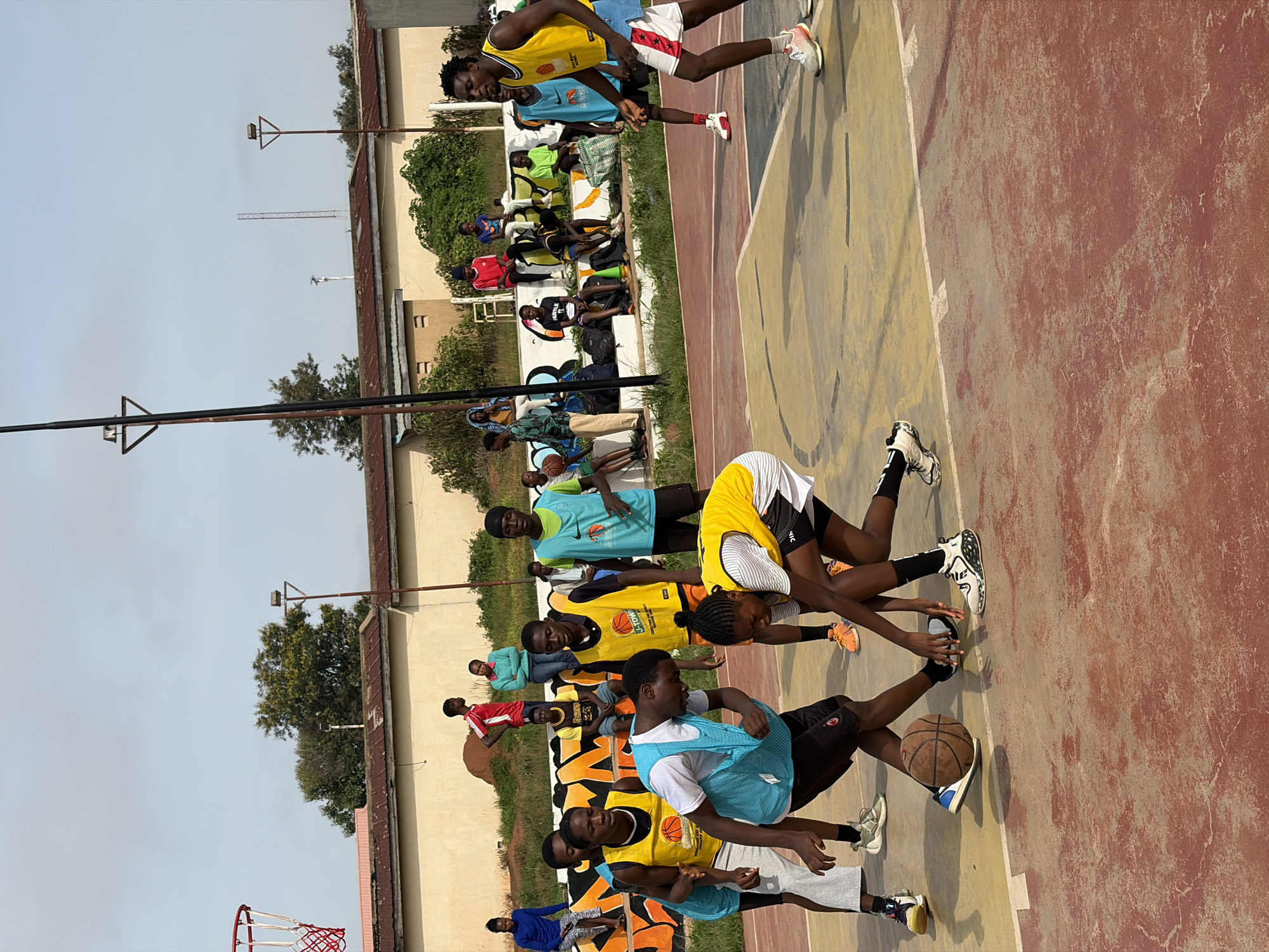 An outdoor basketball court in Jos, Nigeria, is depicted with a group of young people playing a game. The court has a worn, reddish-brown surface with yellow painted lines. In the foreground, two individuals are engaged in a play, one dribbling a basketball while the other attempts to defend. Several other participants are scattered across the court, some actively playing and others observing. In the background, a beige building with a corrugated iron roof is visible, and a chain-link fence encloses the area. A basketball hoop with a red net stands to the left of the frame. The sky is overcast.