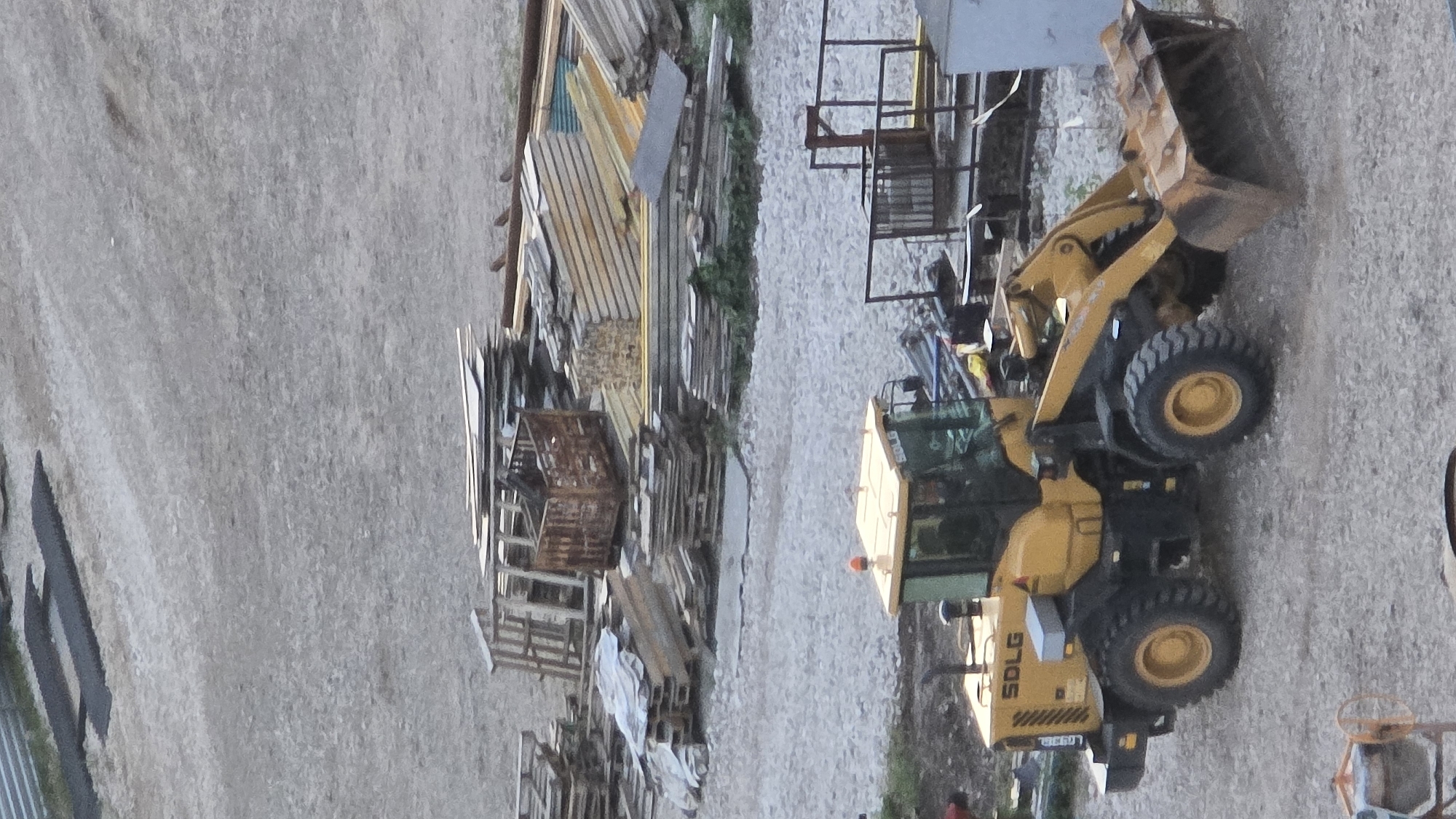 This media file depicts an outdoor scene, likely a construction or industrial yard, given the presence of various materials and heavy machinery. A yellow SDLG wheel loader is the central object, positioned on a gravel surface. The loader has its bucket raised. To the left of the loader, there is a substantial pile of lumber, metal sheeting, and other building materials stacked and organized. Some of these materials are weathered, suggesting they may be in storage or awaiting use. The ground is covered in small gray gravel, and there are hints of greenery in the background, possibly grass or low-lying vegetation.
The image appears to be taken during daylight, with the lighting suggesting it could be late afternoon or early evening, as there are some shadows. The weather seems to be clear. The overall atmosphere is one of an active worksite. A small orange light is visible on top of the loader's cabin, and the text "SDLG" is clearly legible on the side of the machine. There is also a license plate visible on the rear of the loader, though the text is too blurry to be read. A person is partially visible in the lower left corner, wearing dark clothing. Another person is barely visible in the lower right, near some machinery that resembles a concrete mixer. The scene is oriented sideways in the image.