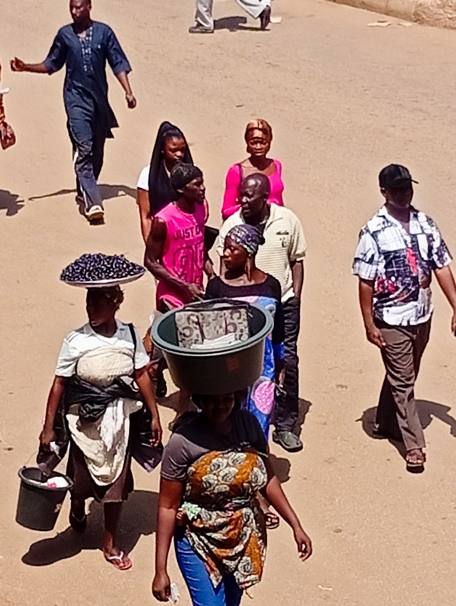 The image depicts an outdoor street scene in Bukuru, Nigeria. A group of people are walking on a sandy or dirt road. In the foreground, a woman is walking from left to right, carrying a dark bucket in her left hand. She is wearing a white and grey striped shirt, a dark skirt, and white socks with red flip-flops. On her head, she carries a large platter piled high with dark, round fruits, likely berries or small plums. Behind her, a person in a pink tank top with the inscription "JUST DO IT 96" is walking away from the viewer. To the right of the person in the pink shirt, another person wearing a light-colored, horizontally striped shirt and dark pants is walking. Further to the right, a man wearing a black baseball cap and a patterned shirt with short sleeves is walking towards the right. In the background, a man in a dark blue traditional outfit is walking towards the left, with his right hand raised. Behind him, partially obscured, is another person in a pink top and a woman with dark hair and a headscarf. The environment appears to be a public area, possibly a market or thoroughfare, with the ground showing signs of wear. The lighting suggests it is daytime, with distinct shadows cast by the individuals.