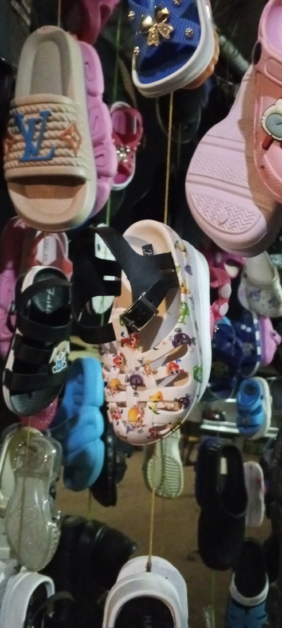 The image shows a collection of children's shoes hanging from strings, likely for sale. The shoes are various colors and styles, including sandals, slides, and crocs. One pair of slides has a design that mimics the Louis Vuitton logo. Another pair of sandals has a black strap. A pair of crocs is patterned with cartoon characters. The background is blurry and dark, but the setting is suggested to be a marketplace or a street vendor stand in Sabon Tasha, Nigeria.