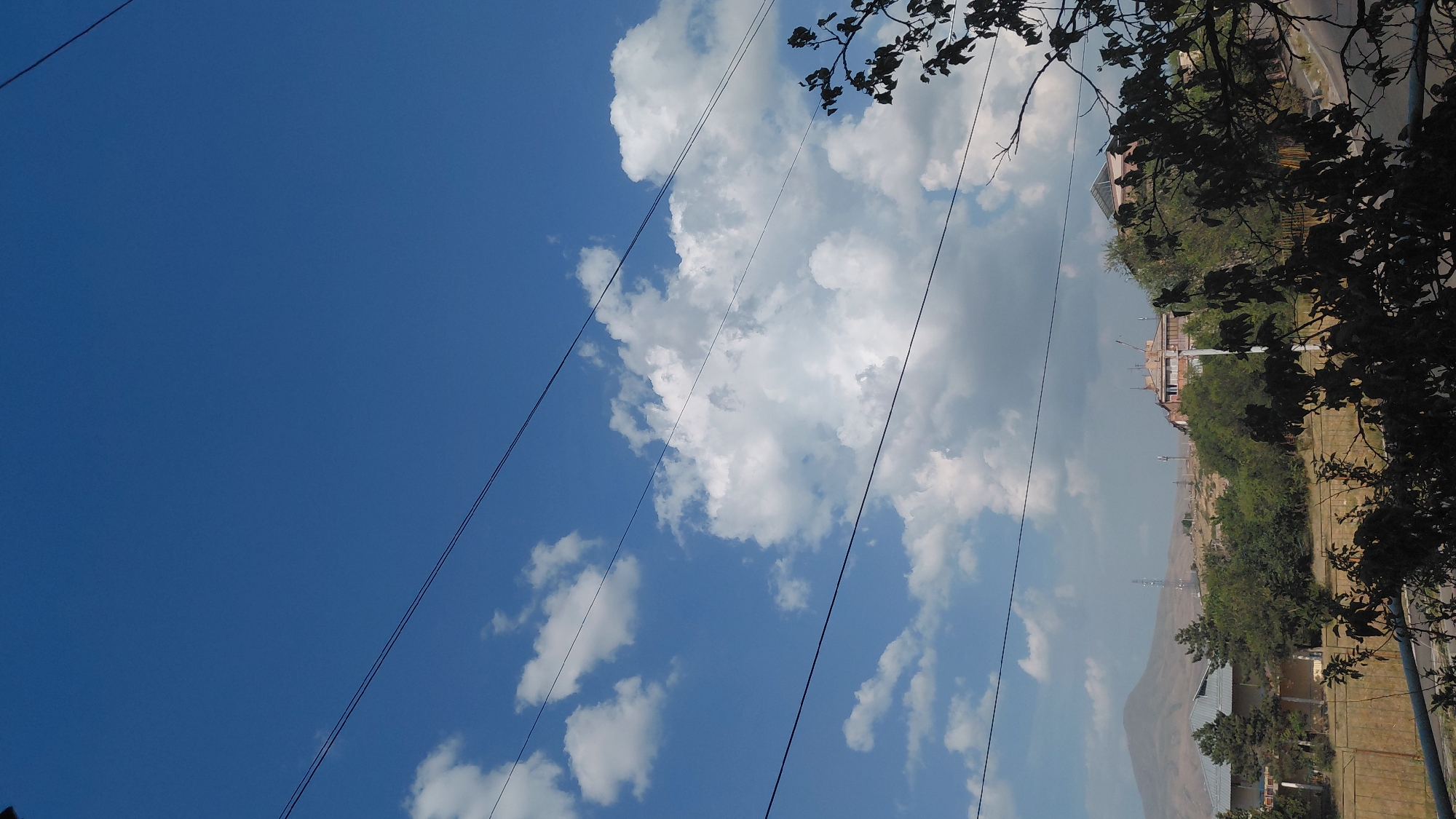 The image shows a view of the city of Abovyan, Armenia, on a clear day with scattered clouds. In the foreground, there are power lines stretching across the frame. Below the power lines, a landscape is visible with trees and buildings. One building, appearing to be a residential structure or a small complex, is situated on a hill and has a light-colored facade with balconies. In the background, a mountain range can be seen, and several communication towers are scattered across the terrain. The overall scene depicts a somewhat rural or suburban area with evidence of infrastructure and natural elements.
