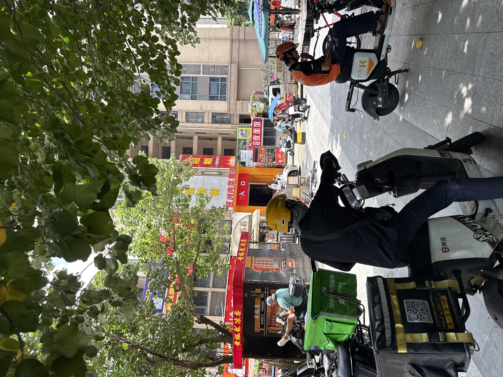 In Haizhu District, China, a street scene unfolds under the dappled shade of lush green trees. The foreground is dominated by several parked electric scooters, some equipped with large, insulated delivery bags. One scooter in the immediate foreground has a black and yellow delivery box displaying a QR code. To the right, a person in an orange vest and black pants sits on an electric scooter, looking down at their phone. Another person, wearing a black shirt and a yellow helmet, is seated on a scooter further to the left, also appearing to use their phone. In the background, a row of shops and buildings line the street. One shop, brightly colored in red and white, displays Chinese characters. People are visible in the distance, some walking and others on scooters, suggesting a busy urban environment. The overall impression is one of everyday street life and commerce in a Chinese city.