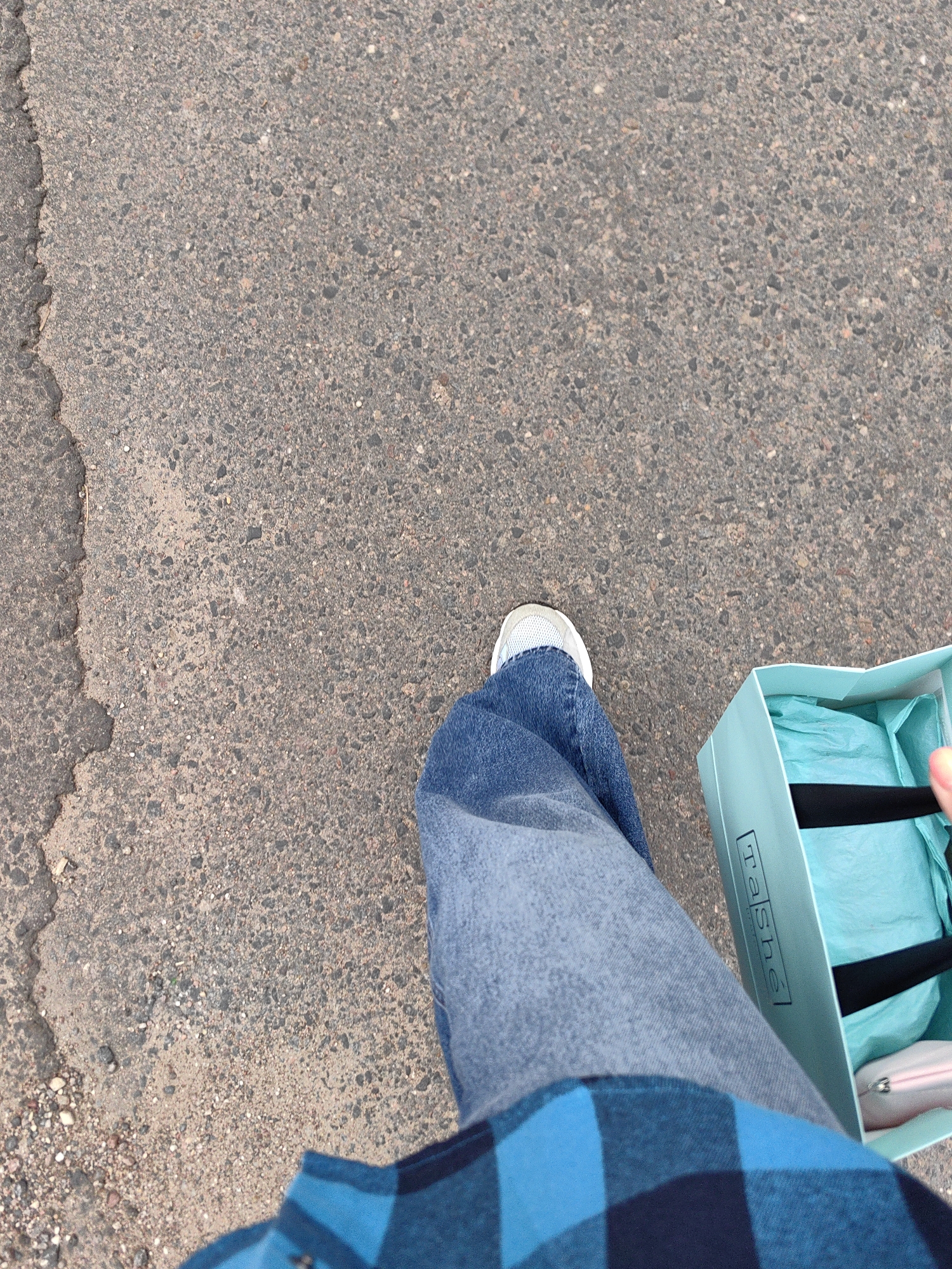 A first-person perspective shot captures a person walking on a paved surface, likely a sidewalk or road, in Fanipal, Belarus. The ground is a rough, grey asphalt with visible cracks and small stones. The person is wearing blue jeans and a blue and black checkered shirt. They are also wearing white sneakers. In their right hand, they are carrying a light blue shopping bag with black straps and a logo that reads "Tashé". Inside the bag, light blue tissue paper is visible, along with a light pink object, possibly a wallet or small purse. The scene appears to be outdoors, with natural light illuminating the area.