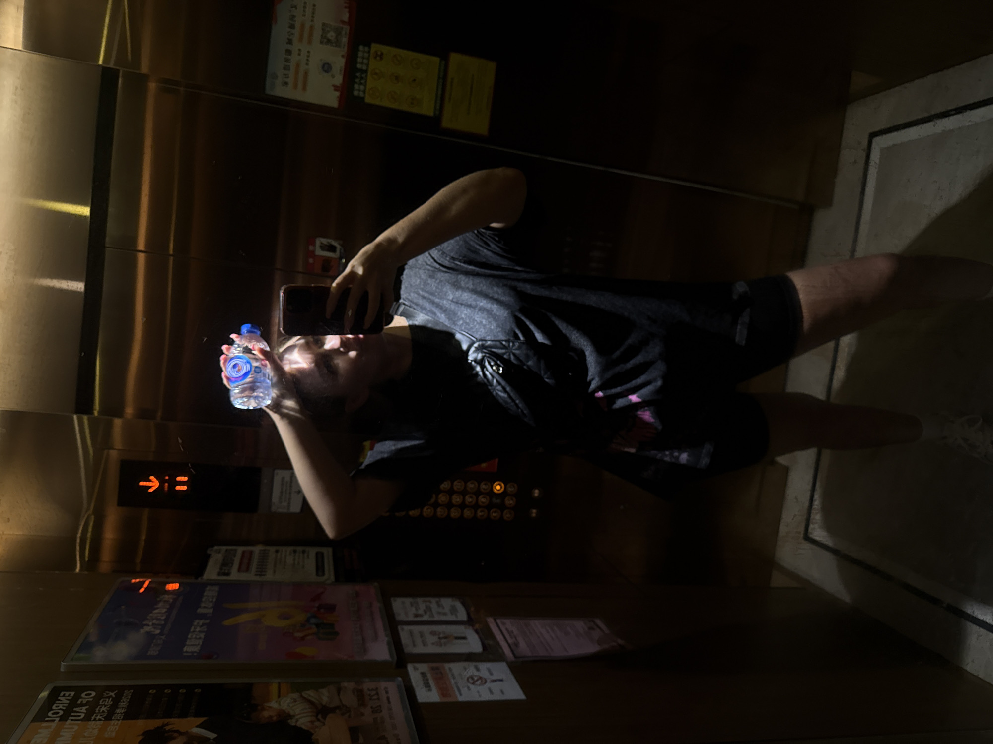 An image, oriented ninety degrees counter-clockwise, depicts a person taking a mirror selfie inside a dimly lit elevator in Yiwu, China. The metallic walls of the elevator are highly reflective, showcasing the person's full-body reflection.
The subject, appearing to be a young adult, wears a dark t-shirt and dark shorts, complemented by white sneakers. Their left arm is raised, holding a dark-colored smartphone to capture the reflection. In their right hand, they hold a small, clear plastic water bottle with a blue cap and a blue-and-white label, which appears to emit a soft blue glow, possibly from internal illumination or strong light reflection. The person's face is partially obscured by the phone and the angle, but their head is tilted slightly upwards.
The elevator's interior features warm-toned, possibly brass or bronze, paneling. An illuminated orange arrow pointing upwards (↑) is visible on a panel above a set of circular elevator buttons, one of which glows orange. Below the buttons and to the left, multiple signs and posters are affixed to the wall. These include white and yellow-bordered rectangular signs displaying Chinese characters, some with icons such as a QR code or "no smoking" symbols. One larger poster on the lower left has both Chinese and partially visible English text, reading "ENROLL" and "OF AUTUM," suggesting an advertisement for an educational institution or event. The overall lighting is subdued, giving the scene an evening or nighttime ambiance.