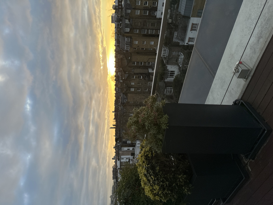 An elevated view looks onto a city bathed in the golden light of a sunrise or sunset. Dense clusters of buildings line the horizon, transitioning into a sky filled with layered clouds. In the foreground, planters filled with green foliage accent the scene. The edge of a modern balcony, featuring glass and dark wood, frames the cityscape.