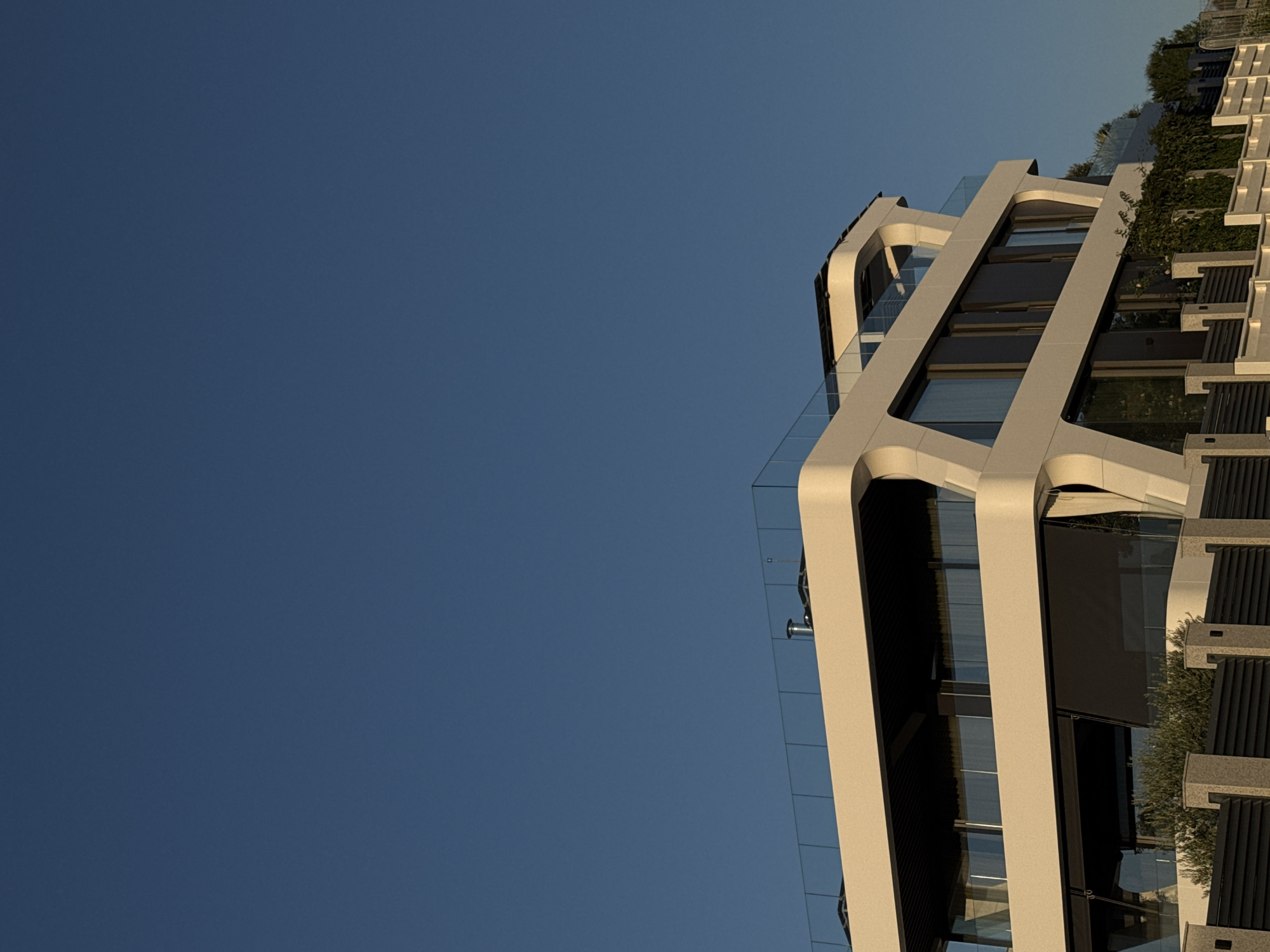 An outdoor, eye-level, medium shot captures a modern, multi-story building against a clear, deep blue sky. The building's exterior is primarily white with a curved, futuristic design. Large, dark-tinted windows reflect the sky. A glass railing encircles the top level, and solar panels are visible on the roof. The lower portion of the building features a black retractable awning and greenery climbing the exterior wall. In the foreground, a dark metal fence with stone pillars separates the property from the viewer. The scene appears to be bathed in the warm light of late afternoon or early morning. The location is specified as Lozenets, Bulgaria.