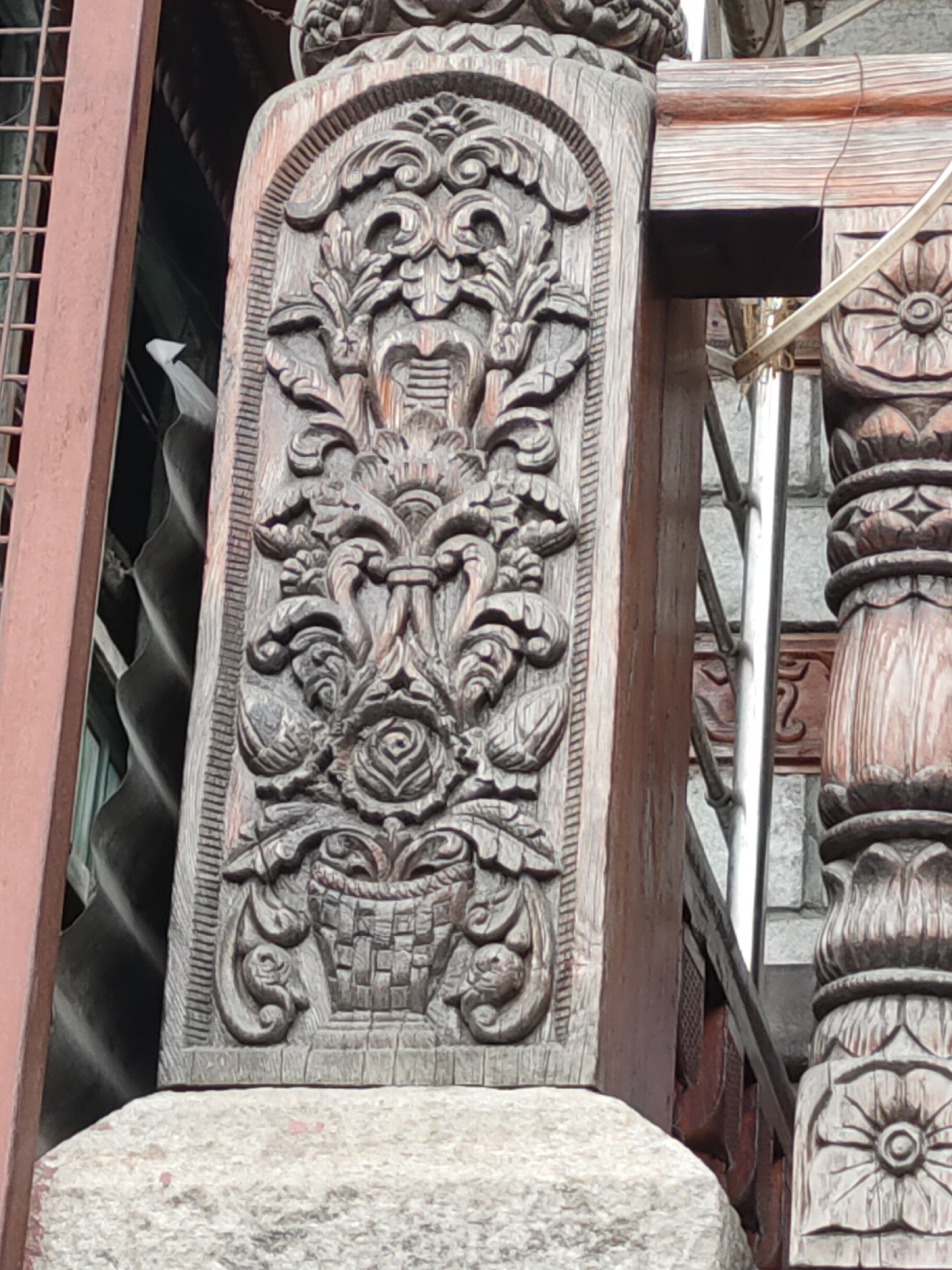 The image shows a close-up of intricate wood carvings on a structural element. The carving features a central motif resembling a basket filled with floral elements and leaves, bordered by ornamental scrollwork. The wood appears weathered and aged, with visible grain and a dark brown stain. To the left, a reddish-brown metal railing or frame is visible, partially obscuring a corrugated metal surface. To the right, another carved wooden post with floral designs and a lighter wood grain is partially visible, along with a silver-colored metal pipe. The carving is set against a background that includes stone or concrete elements at the bottom and a textured gray surface behind the metal pipe. The scene suggests an architectural detail from an outdoor setting, potentially in a location with decorative craftsmanship.