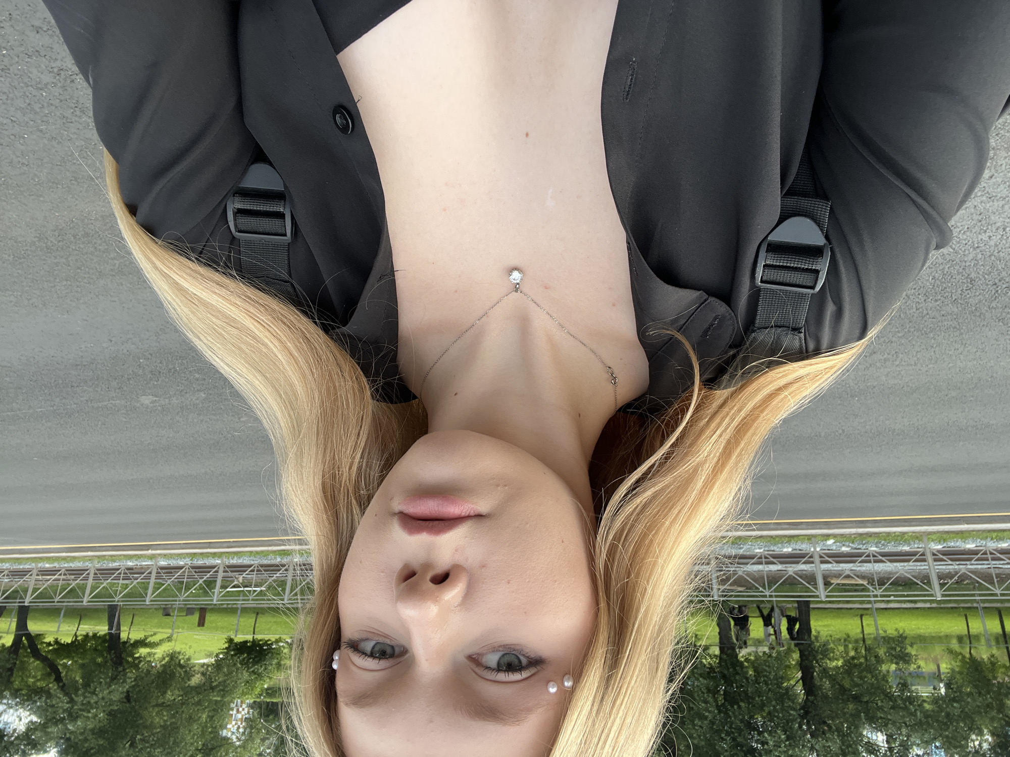 A young woman with long blonde hair and light blue eyes is photographed from the chest up, facing the camera. She is wearing a black collared shirt, open to reveal a black top and a silver necklace with a clear pendant. Small pearl-like embellishments are applied to her eyelids. The background shows a street, a pedestrian walkway with a metal fence, railroad tracks, a grassy park area with trees, and buildings in the distance. The scene appears to be outdoors, likely in an urban setting such as Perm, Russia. The lighting suggests it is daytime.