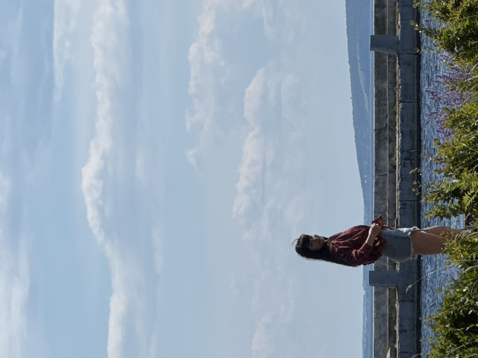 An outdoor, medium shot captures a young woman with long, dark hair, wearing a maroon and white checkered long-sleeved shirt and light blue denim shorts. She stands on a concrete pathway, her body turned to her left as she looks back over her shoulder towards the camera with a slight smile. The pathway appears to be a pier or sea wall. Behind her, a body of water is visible, stretching towards a distant, hazy coastline. The sky above is a bright blue, dotted with scattered, fluffy white clouds. In the foreground, lush green foliage with hints of purple flowers partially obscures the lower portion of the image. The lighting suggests a bright, sunny day. The scene is located in an unspecified outdoor location.