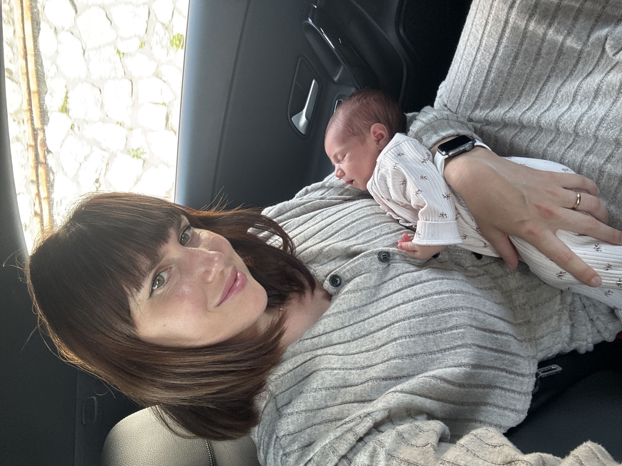 A medium shot captures a woman holding a baby in a vehicle. The woman, with brown hair and bangs, smiles toward the camera. She wears a striped gray sweater. The baby, dressed in white patterned clothing, is asleep on her chest. The woman wears a watch and a gold ring. The interior of the car is visible, with the door handle in view. Outside the car window, a stone wall is partially visible. The photograph was taken in Nettuno, Italy.