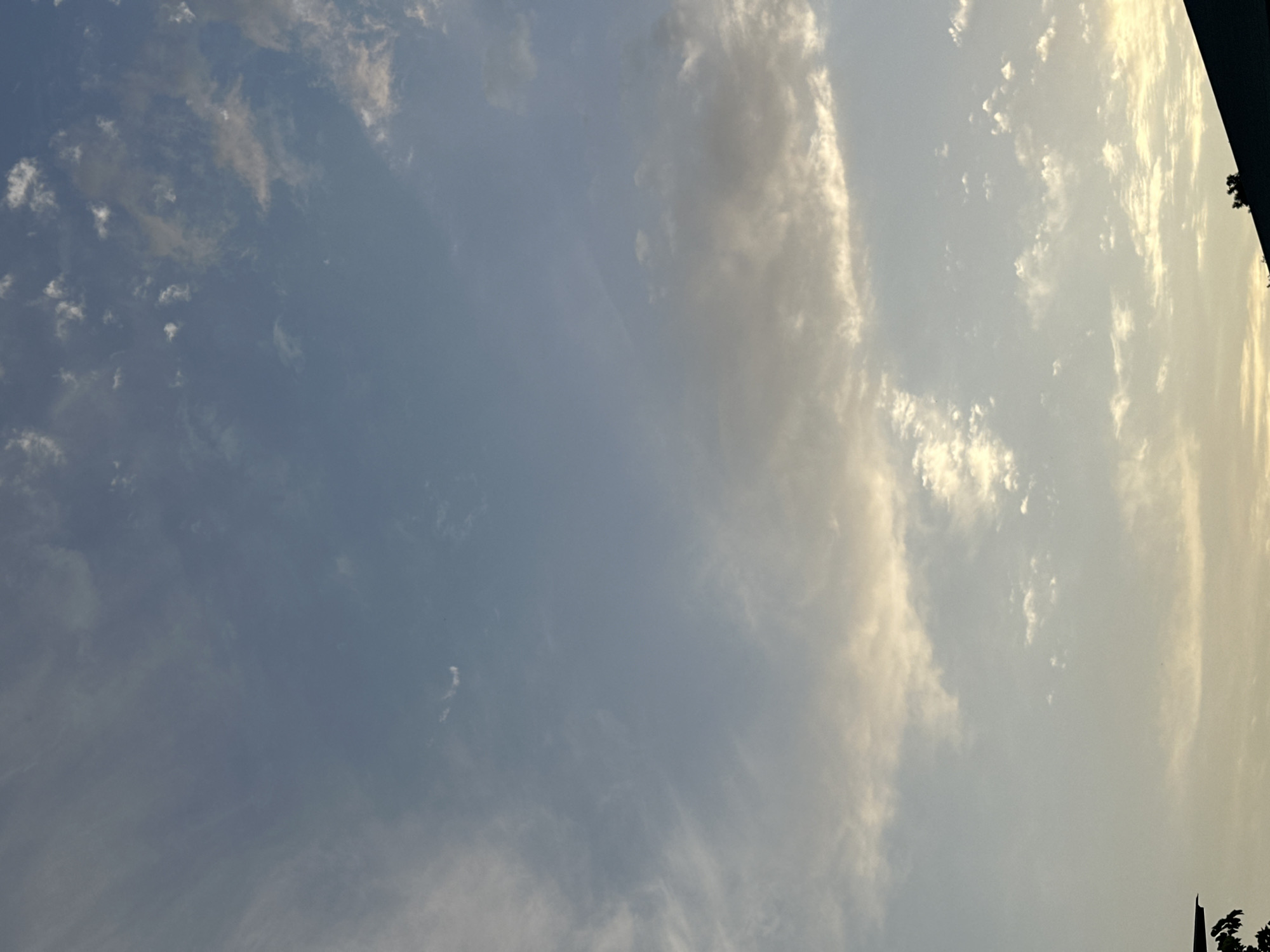 The image depicts a clear sky with wispy clouds and a soft, diffused light, suggesting either early morning or late afternoon. The top portion of the frame is dominated by a pale blue sky with scattered, thin clouds that are translucent and appear to be streaked. Towards the middle and bottom, the clouds become more defined and are illuminated with a warm, golden hue, indicating the direction of the sun. The very bottom of the image shows the dark silhouette of a rooftop and some foliage, indicating an outdoor setting in a built-up area. The scene is devoid of any people or specific actions. The overall impression is one of a peaceful sky. The location in Sasbaga, Uzbekistan, as per the prompt, cannot be visually confirmed from the image content.