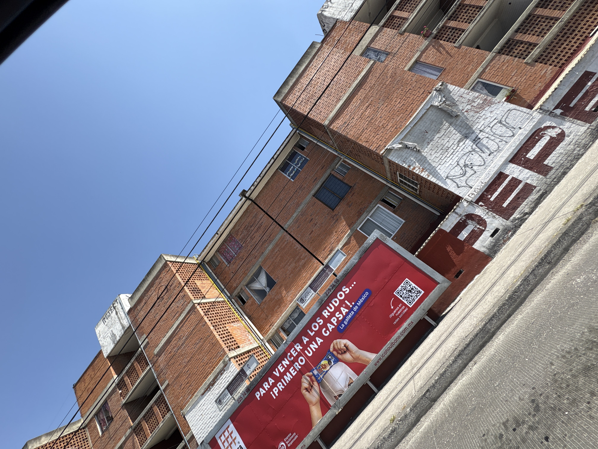 An urban scene in Puebla, Mexico, captured with a tilted perspective, looking upwards at multi-story residential buildings and a clear blue sky.
The foreground features a concrete sidewalk or curb on the lower left. Directly behind it, a white-painted wall exhibits large, faded red lettering spelling "PEPE," partially obscured by black spray-paint graffiti. Above this wall, a large, rectangular red billboard is prominently displayed. The billboard advertises "GAPSA!" cookies, featuring a graphic of two hands holding a blue-packaged cookie. Spanish text reads: "PARA VENCER A LOS RUDOS... ¡PRIMERO UNA GAPSA!" (To defeat the tough guys... First a Gapsa!), with the subtitle "La galleta de México" (The cookie of Mexico). A website, "www.iconourbano.com.mx," and a QR code for social media are visible on the billboard, along with a "Socially Responsible Company" logo.
Behind and above the billboard are multi-story buildings constructed primarily of red brick. These structures feature numerous windows, some with white frames and visible interiors, and balconies with concrete ledges and open brickwork patterns (celosías). Portions of the building facades are painted white, displaying wear and additional graffiti tags in black. Black utility lines stretch across the frame, connecting to the buildings. A concrete rooftop structure, also marked with graffiti, is visible at the apex of one building section on the right. A phone number, "225 74 98," is visible in white brick sections above the billboard. The upper right portion of the image is dominated by a clear, bright blue sky, indicating daylight conditions. No individuals are visible in the scene.