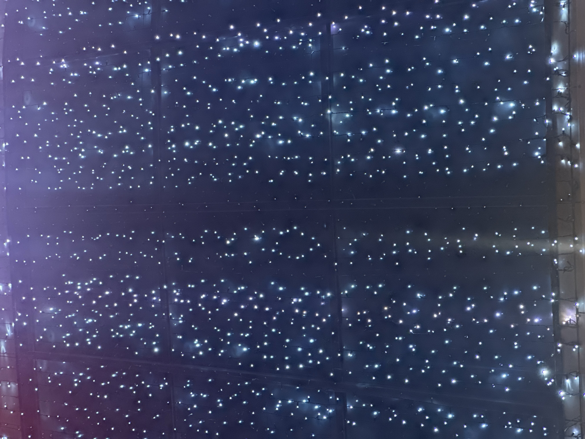 The image captures a dense overhead display of numerous small, bright, cool-toned lights set against a dark, deep blue-purple background. The lights are arranged in an organized grid-like pattern, forming distinct horizontal rows and vertical columns. Thin, dark wires connecting the individual lights are faintly visible against the darker backdrop. The background itself appears to be a solid surface, possibly a ceiling or a large panel, with subtle structural lines or divisions running through it, which support or are part of the light installation. The overall effect creates a sparkling, ethereal atmosphere, reminiscent of a starry night sky or festive decorations. There is no human activity, visible text, or discernible weather conditions. The scene is illuminated at night or in a dark interior setting, likely part of an architectural or decorative installation in Stavropol, Russia.