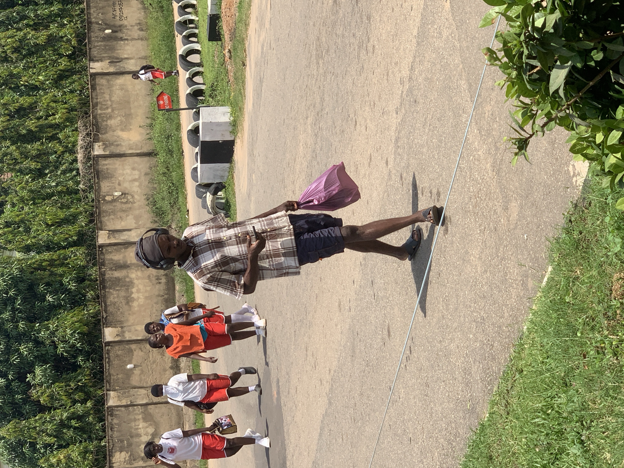 In the city of Abuja, Nigeria, a group of five young people, four females and one male, are walking on a paved road under a clear sky. The four females are dressed in matching white t-shirts and red shorts, with two of them carrying bags. The male, walking in the foreground, is wearing a checkered shirt, dark shorts, and headphones, and is looking at his phone while carrying a pink bag. In the background, a concrete wall runs along the side of the road, with lush green trees behind it. Further down the road, a "No Parking" sign is visible, along with a line of tires that appear to be part of a makeshift barrier or obstacle course. The scene suggests an outdoor gathering or activity, possibly related to sports or recreation, given the athletic attire of the individuals.