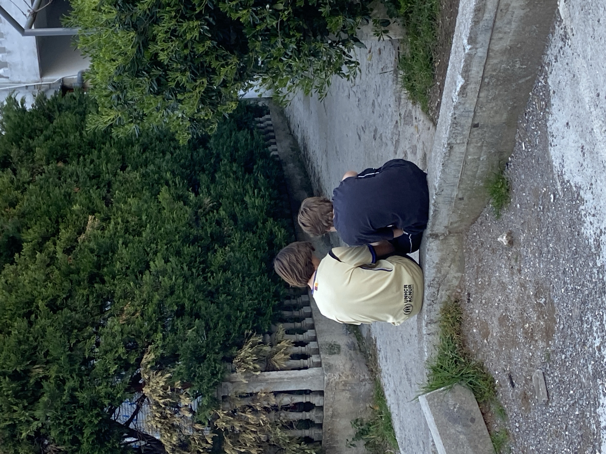 Two children are squatting next to a concrete curb adjacent to a paved walkway. One child is wearing a navy blue shirt and the other is wearing a pale yellow t-shirt with the UNHCR logo. The walkway is lined by a low stone balustrade and lush green bushes, typical of the urban landscape in Budva, Montenegro. The paved walkway stretches into the distance with houses partially visible in the background.