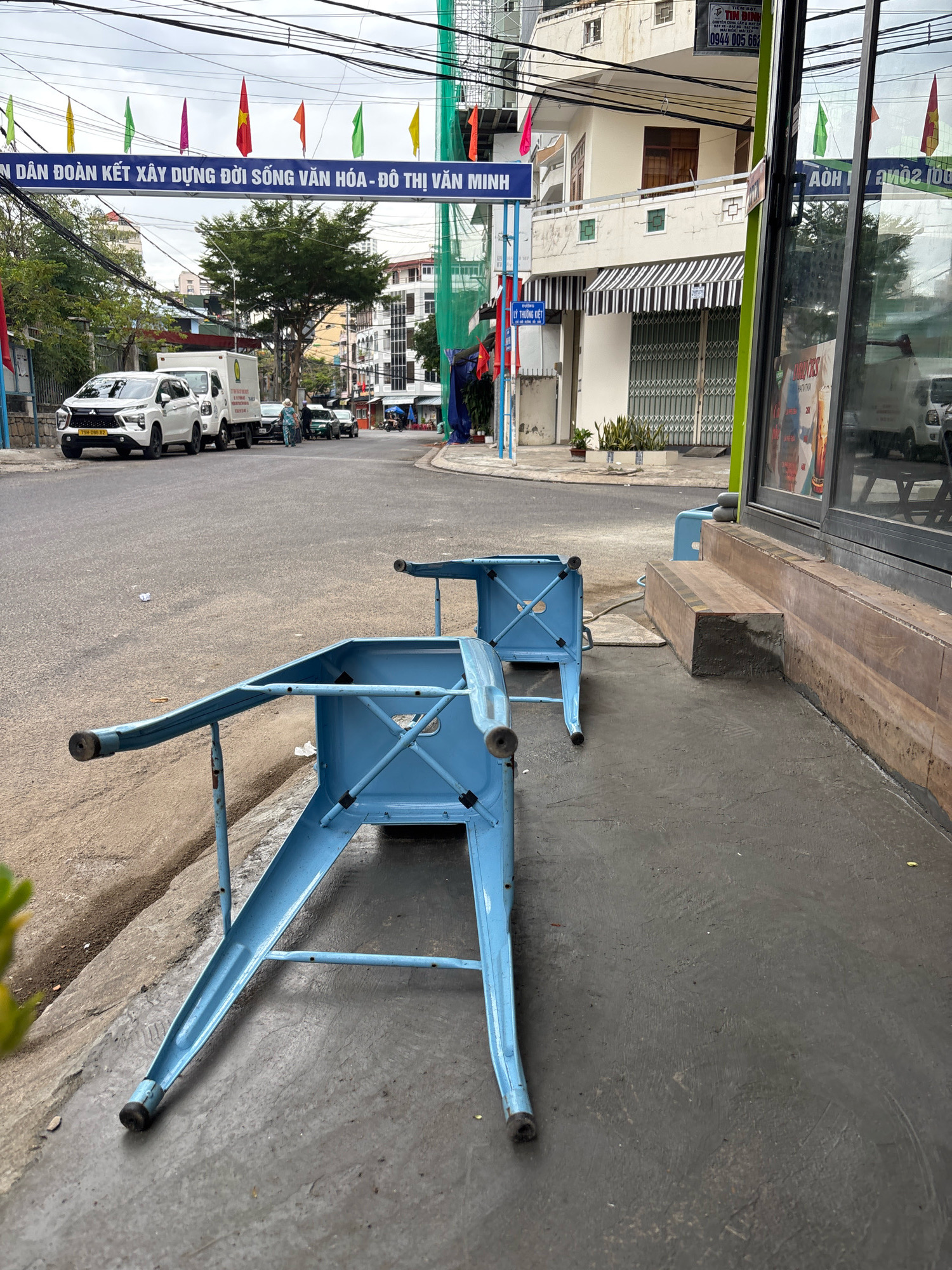 The image depicts a street scene in Nha Trang, Vietnam, under an overcast sky. In the foreground, two light blue metal chairs are overturned on a dark grey paved sidewalk. The chairs are simple in design, appearing to be outdoor cafe seating. To the left of the chairs, a concrete curb separates the sidewalk from a paved street. Several cars are parked or driving on the street, including a white SUV with a license plate ending in 87. Further down the street, buildings line both sides, with one building featuring a black and white striped awning over a closed storefront. Banners with Vietnamese text and flags of various colors are strung across the street and on poles. The overall environment suggests a commercial or residential area within the city. The absence of people actively engaged in the scene, beyond those in vehicles, and the overturned chairs might imply a closure or a break in activity.