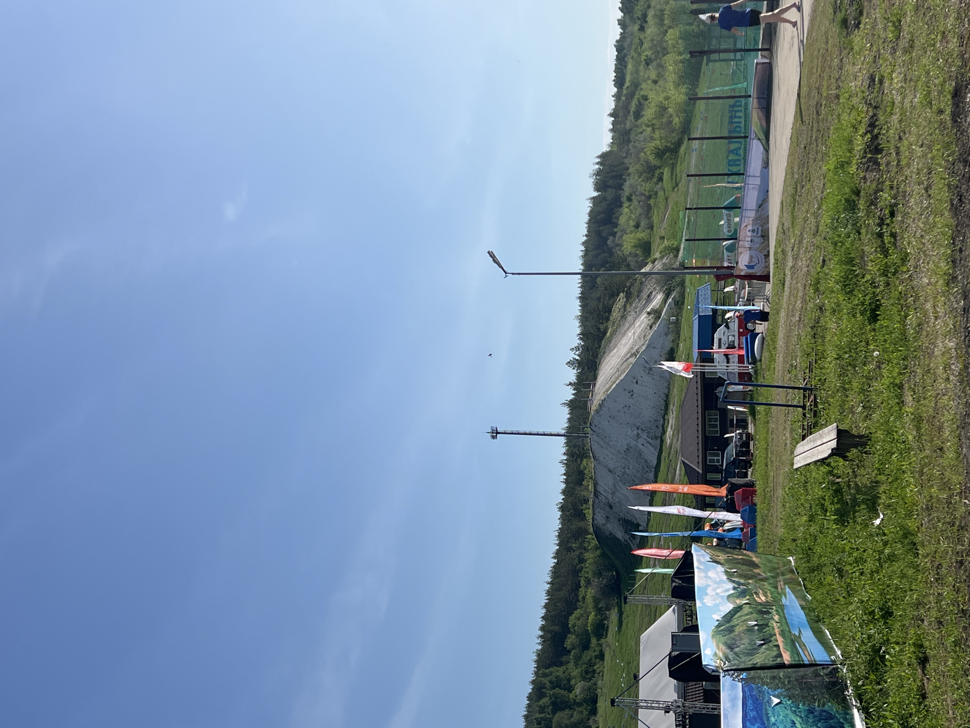 The image captures a landscape view, likely in Khvalynsk, Russia, given the presence of a ski jump hill. The scene includes a ski jump hill covered in white material, possibly snow or a protective covering. Trees surround the hill, indicating a natural environment. In the foreground, there is a grassy area with some flags, tents, and staging equipment. People can be seen in the background, near the top of the ski jump hill. The sky is blue with few clouds. A light pole and communication tower rise into the air.