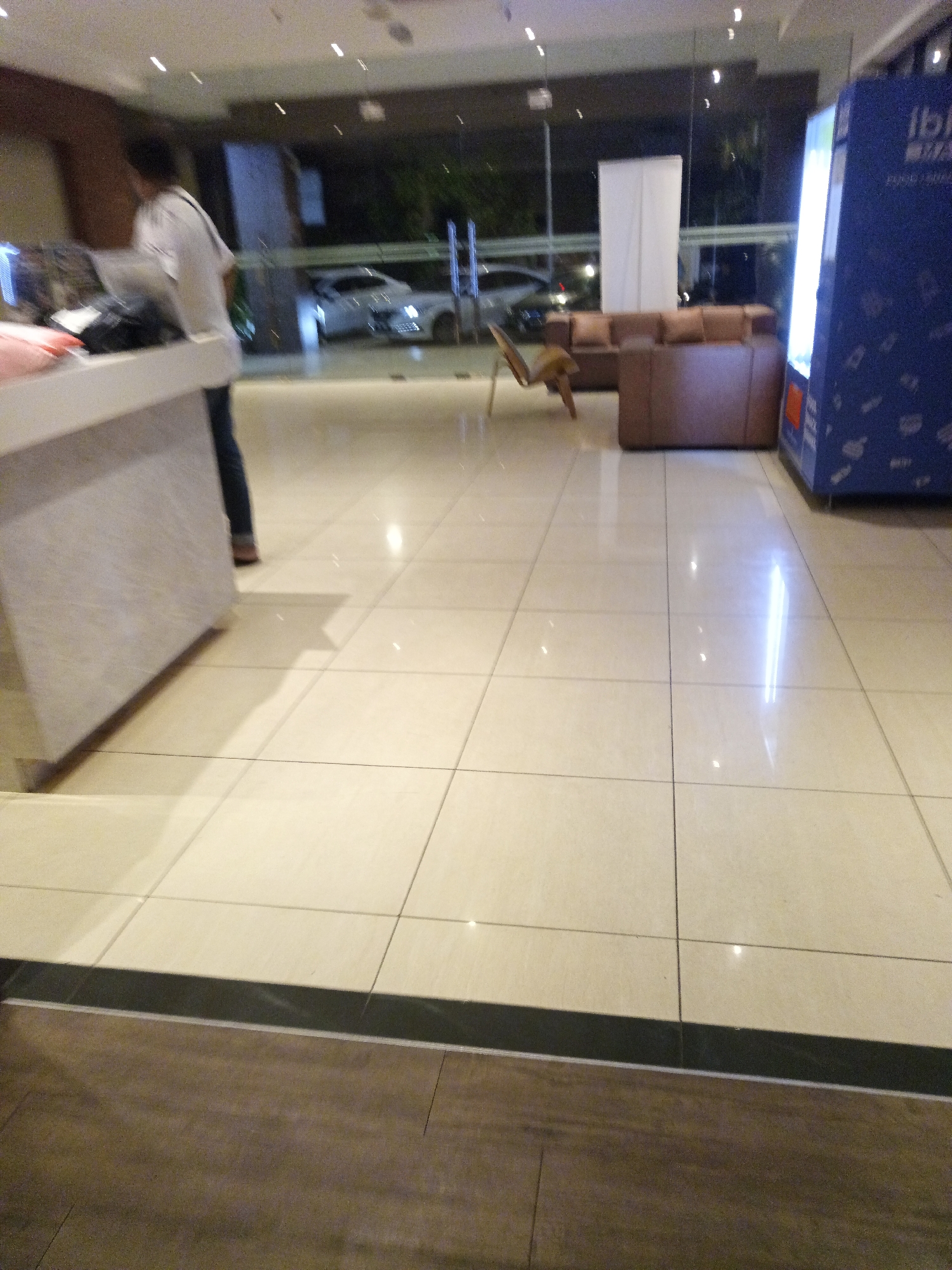 The image depicts the interior of a hotel lobby in Kuala Lumpur, Malaysia. In the foreground, a person wearing a white shirt and dark pants stands behind a white counter. The lobby features a polished tiled floor that reflects the ambient lighting. Through a glass partition, two cars, a white sedan and a darker SUV, are visible parked outside. Further inside the lobby, a brown leather sofa and a single chair are arranged in a seating area. To the right, a tall, blue display with the "ibis" logo is prominently featured. The overall scene suggests a typical hotel reception area.