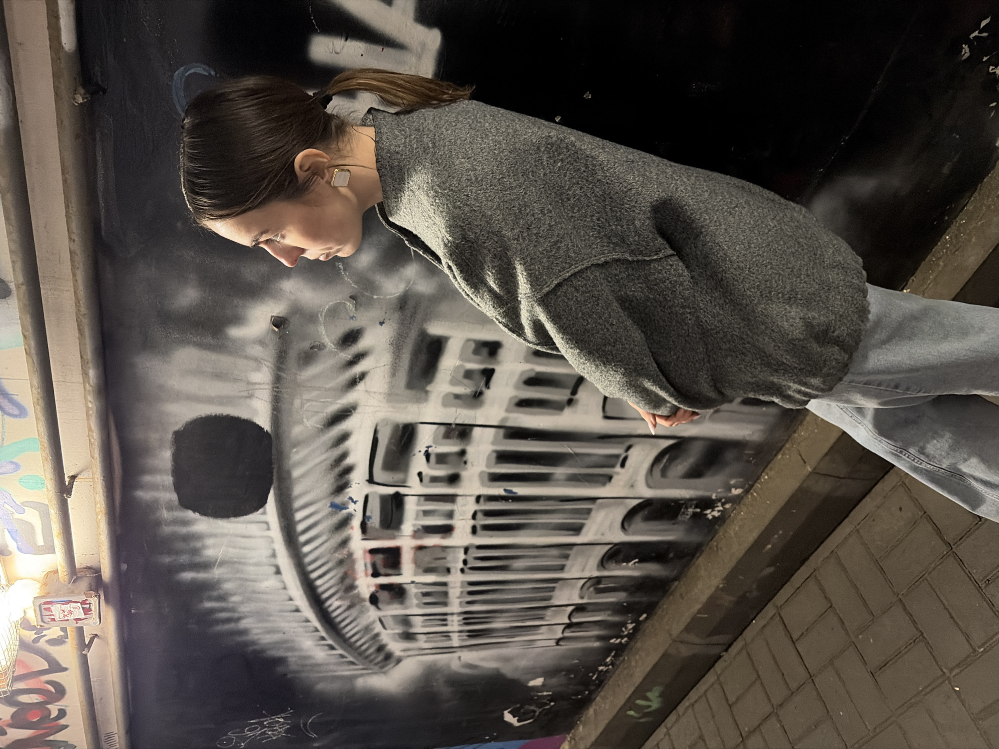 A young woman with dark hair pulled back in a ponytail stands in an underpass in Izhevsk, Russia. She is wearing a gray sweater and jeans. She is looking down at a wall covered in graffiti, including an image that appears to be the facade of a building. The underpass is dimly lit. The wall has some electrical components on the left side. The underpass is paved with bricks.