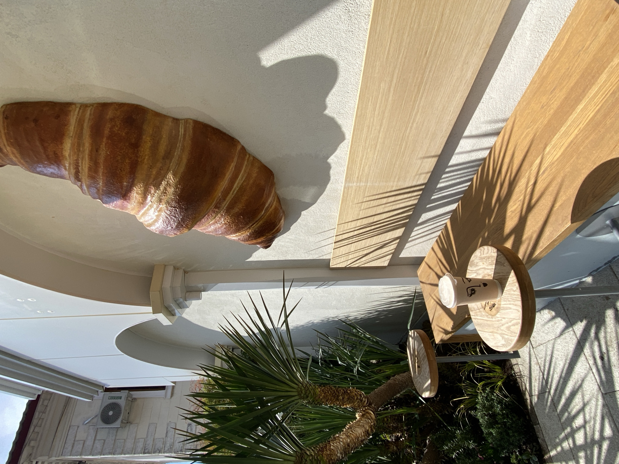 Here's a description of the image:
The image, rotated counter-clockwise, appears to be taken outdoors, likely during daylight, in what looks like a cafe or bakery setting. A large, simulated croissant is mounted on a building exterior. A wooden counter/table is visible, with shadows cast by foliage. A cup, likely containing coffee, rests on a wooden circular support attached to the table. A palm tree is near the building. The outdoor location could be Sochi, Russia.
