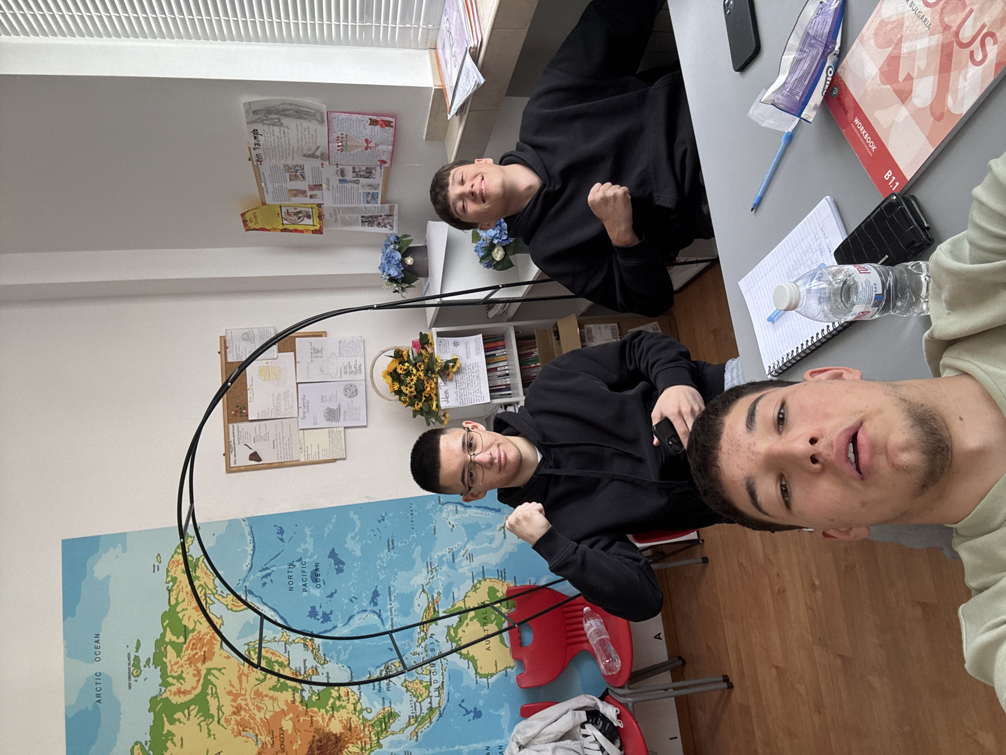 Three young males are captured in a selfie-style photograph within what appears to be a brightly lit classroom or study room. The closest subject, in the foreground, is looking directly at the camera with a relaxed expression and a light goatee, wearing a light-colored top. To his left, another male wearing glasses and a black hoodie smiles at the camera, raising a fist in a playful gesture. Further back and to the right, a third male in a black hoodie also smiles and makes a similar fist gesture.
The room features light-colored walls, windows with horizontal blinds at the top, and a wooden floor. A large world map is prominently displayed on the wall behind the subjects, showing landmasses like Australia, Indonesia, and Mongolia, and oceans like the Arctic and North Pacific. Desks are arranged in the room, with various items visible on one desk including a black smartphone, a blue pen, a partially consumed "OREO" snack package, a black wallet, a notebook, and a "Devin" brand water bottle. A "WORKBOOK B1.1" with "BULGARIA" and "FOCUS" visible on its cover rests on the desk. Shelves filled with books are visible in the background, along with decorative artificial flowers in pots. A cork board on the wall displays several papers, one of which has "Mein Traumjob" (German for "My dream job") written on it.
The scene suggests a casual, perhaps educational, setting during the daytime. The subjects appear cheerful and engaged, possibly posing for a group photo or selfie. The presence of the "BULGARIA" text on the workbook and the "Devin" water bottle, along with the location context, suggest the image was taken in Bulgaria.