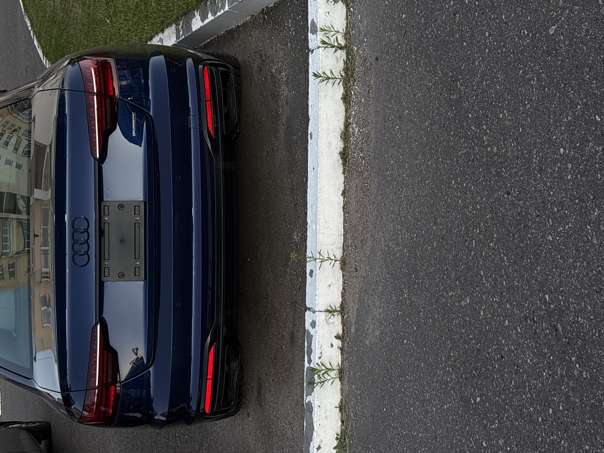 The image shows the rear of a dark blue Audi A4 sedan parked on an asphalt surface. The car has its brake lights illuminated, which appear as thin red horizontal strips. The Audi rings logo is visible on the trunk, along with the "A4" designation and the "quattro" badge. The car is parked next to a white concrete curb, with some sparse green vegetation growing along its base. In the background, a light-colored building with multiple windows can be seen, reflecting in the car's trunk. The scene is static and appears to be a typical urban parking situation. There are no people visible in the image. The location is identified as Vitsebsk, Belarus.