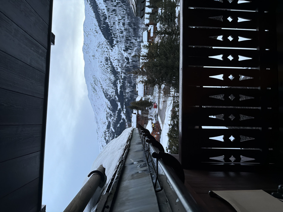 From a balcony, a view of a snow-covered mountain and nearby buildings is framed. A dark wooden door is to the left, with a carved wooden railing on the right. A snow-covered gutter runs along the bottom of the image. The sky is overcast. Buildings are visible in the valley below the mountain.