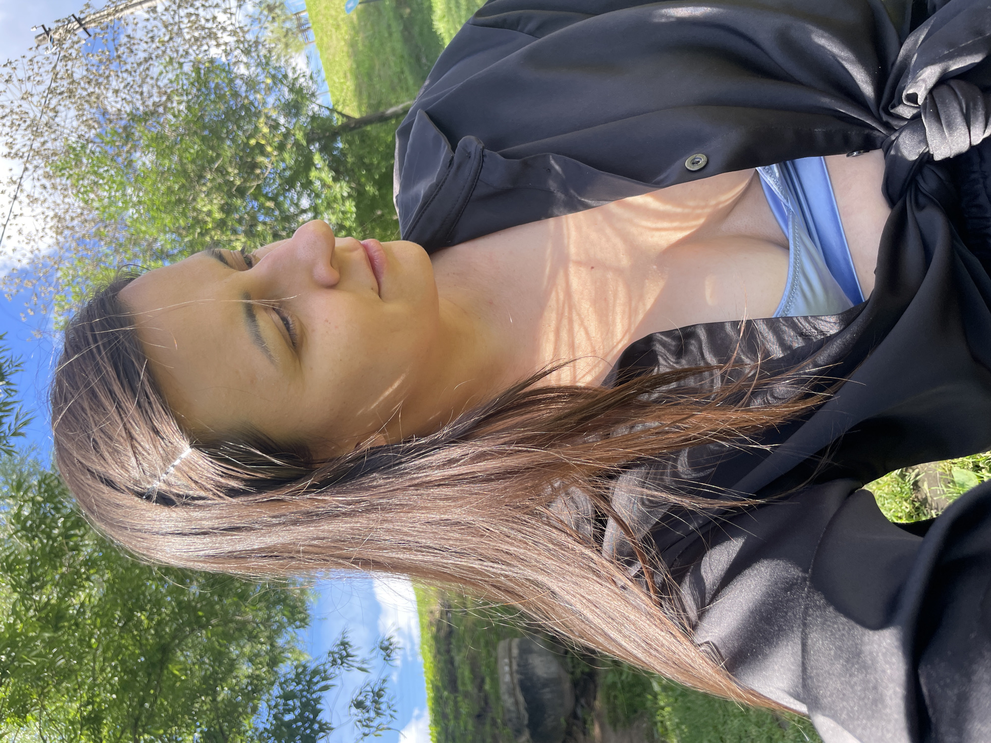The media file is a close-up, vertically oriented photograph featuring a woman with long, brown hair. She is wearing a black jacket, partially open, revealing a light blue garment underneath. Her eyes are closed, and her head is tilted back, suggesting relaxation or enjoyment of the sun. The background is a blur of green foliage and blue sky, indicating an outdoor setting, likely during the day, with good weather. The lighting suggests it is daytime, possibly late morning or early afternoon, with sunlight creating shadows on her skin and clothing. The overall mood is serene and casual. The specific location is Izhevsk, Russia, though no explicit visual cues within the image confirm this. There is no visible text in the image.