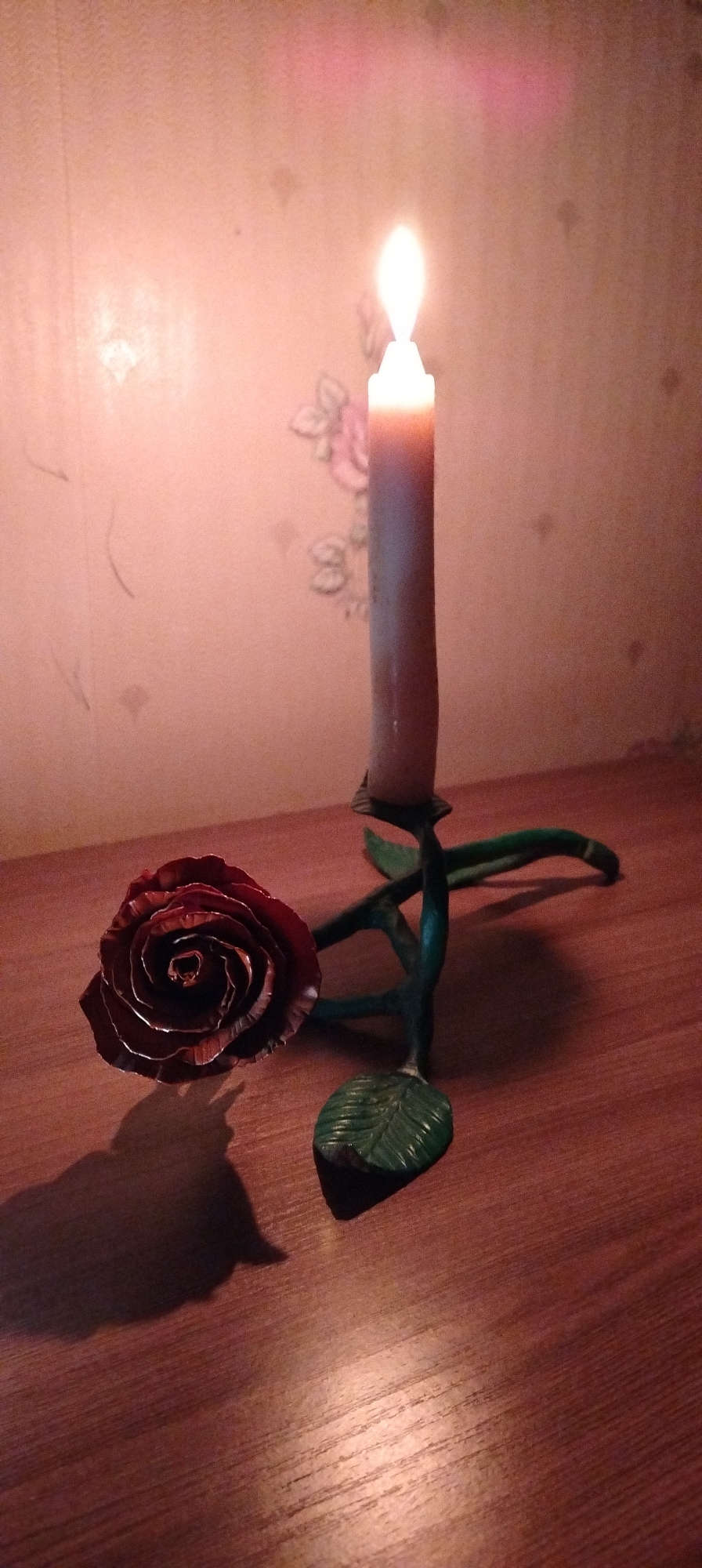 A candle is lit in a metal rose-shaped candle holder on a wooden surface. The walls are off-white with a floral design. The shadow of the rose is prominent on the wooden surface. The location is Зипуново, Russia.